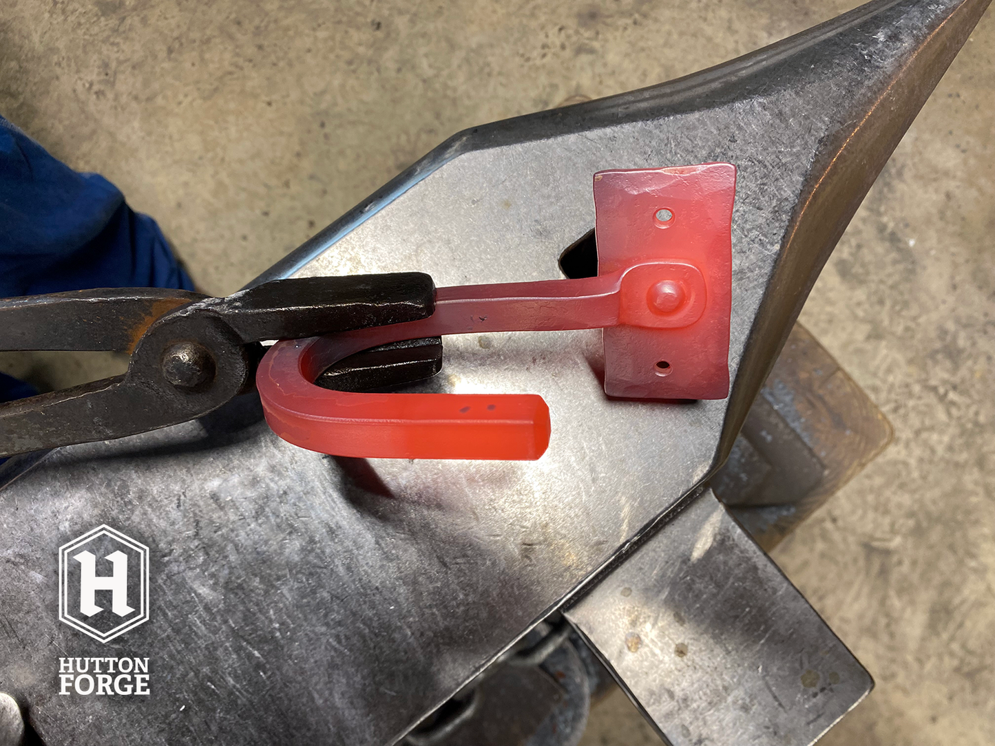 A blacksmith's forge with a glowing red metal gun hook being shaped on an anvil. The hook is held by tongs, and the Hutton Forge logo is visible in the lower left corner.