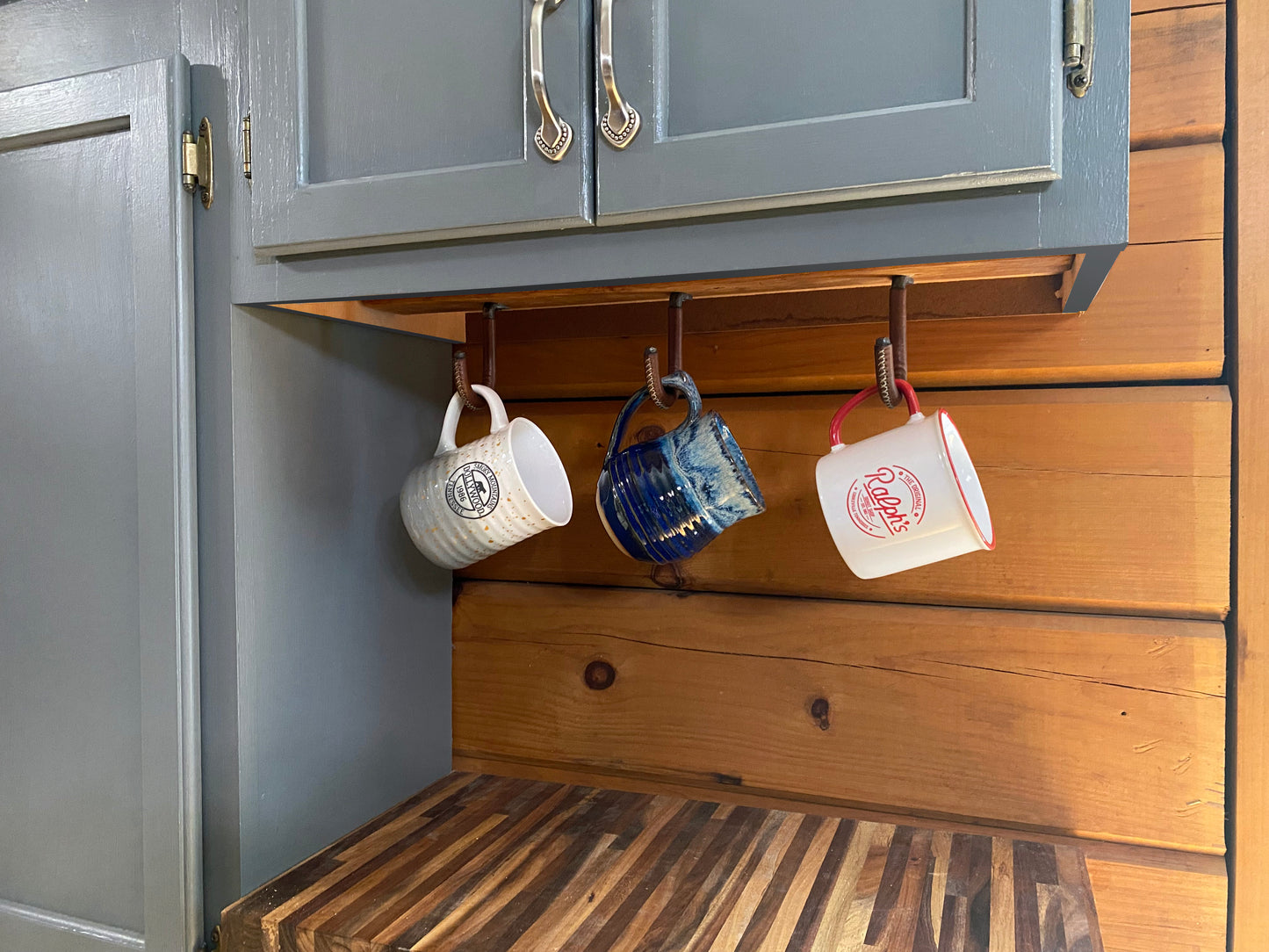 Use multiple hooks to create a unique under cabinet display of your favorite coffee mugs. Three ceramic mugs hanging from rustic leather wrapped metal hooks under kitchen cabinets. Hutton Forge