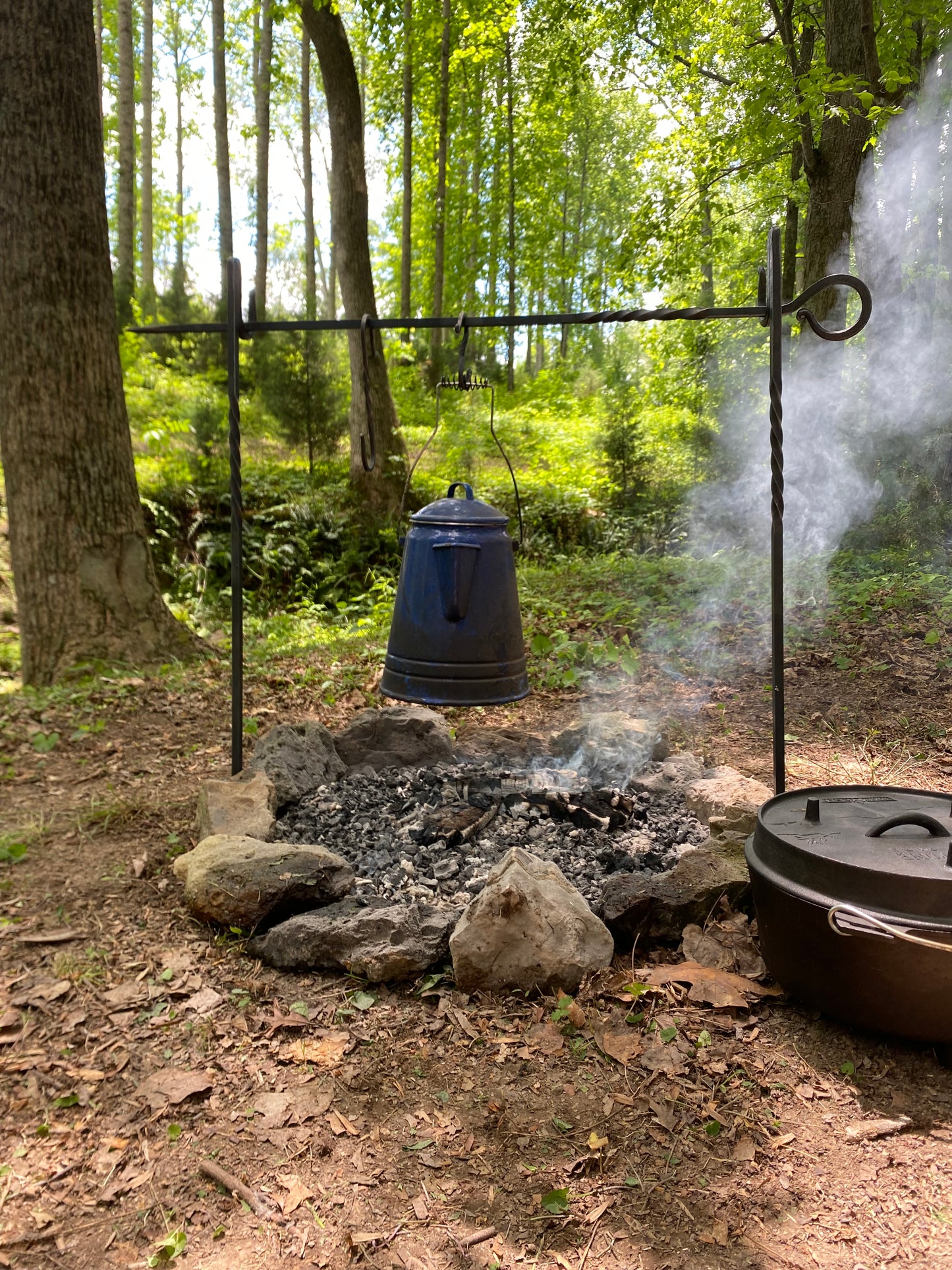 A rustic outdoor cooking setup in a wooded campsite, featuring a dark blue kettle hanging from a black metal tripod over a stone fire pit with smoke rising, alongside a dark brown cooking pot on the right.