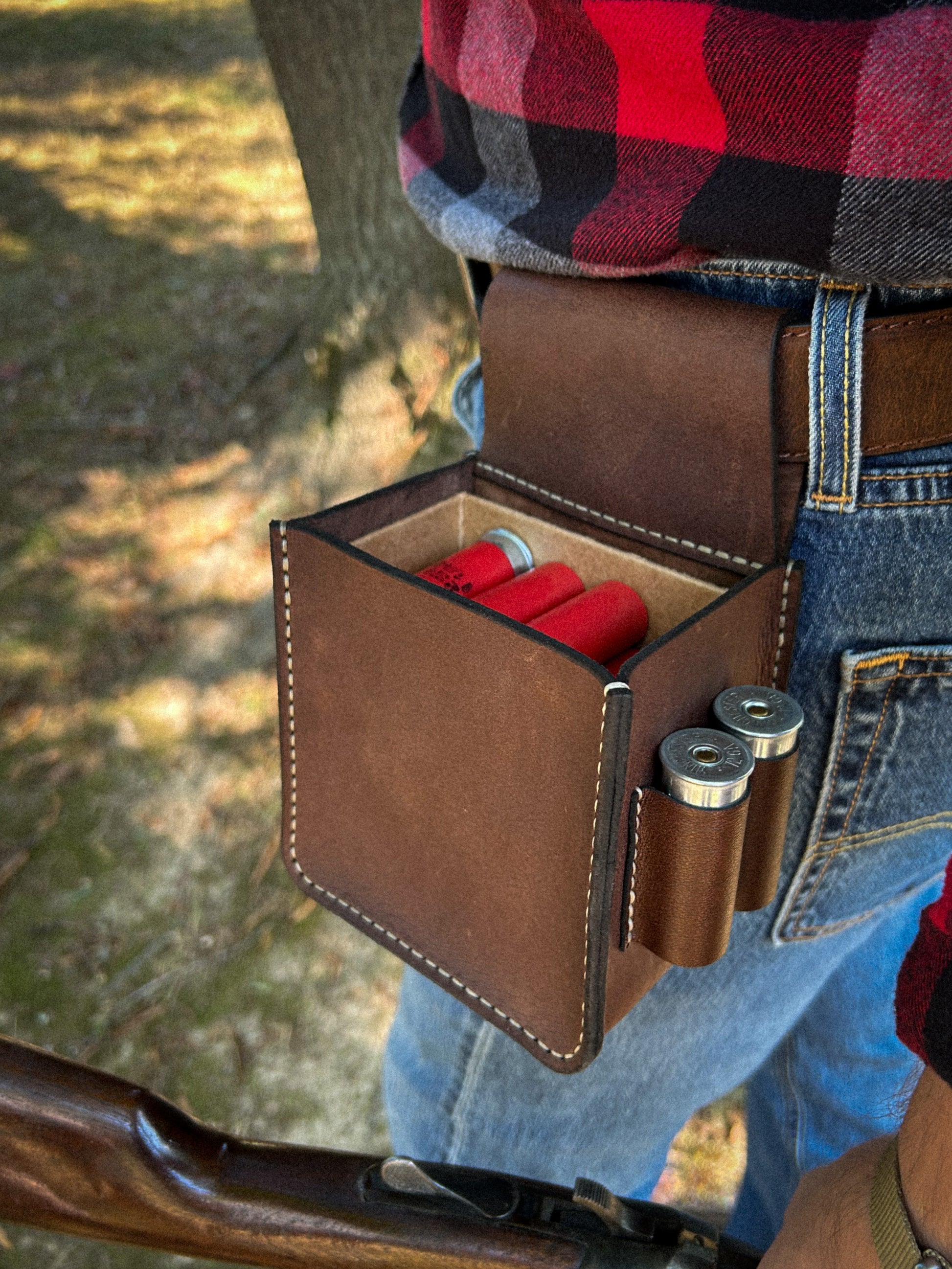 A brown leather shotgun shell pouch worn around a person's waist, holding red shotgun shells, with man holding a gun and wearing a red and black plaid shirt and blue jeans visible in an outdoor setting.