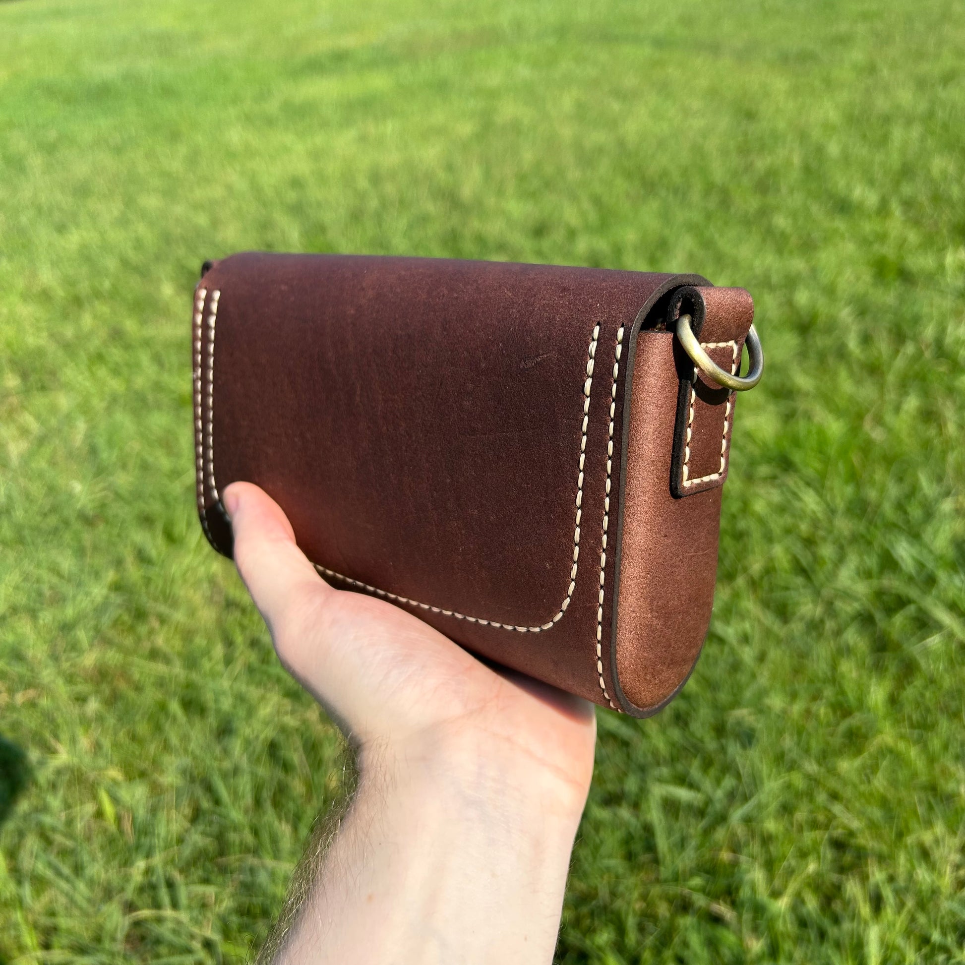 The back of a leather purse with tan stitching and a brass ring, held in a hand against a blurred green grass background.