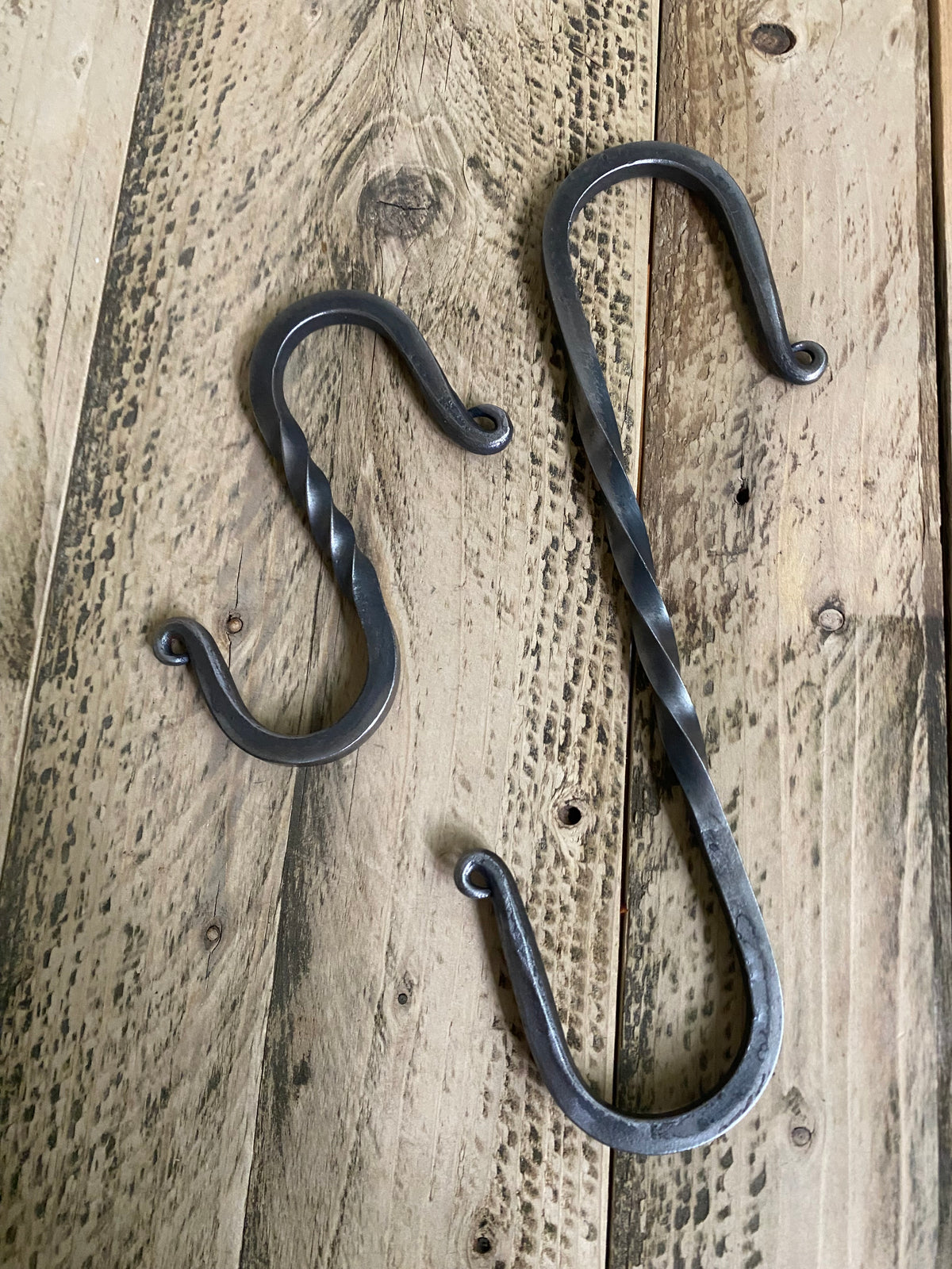 Two hand-forged twisted steel S-hooks side by side, one small and one large, in a blackened finish displayed on a rustic wooden plank surface. Hutton Forge.
