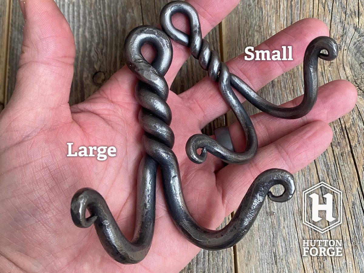 Hand holding a large and small hand-forged twisted double wall hook on rustic wood, labeled Large and Small. Hutton Forge.
