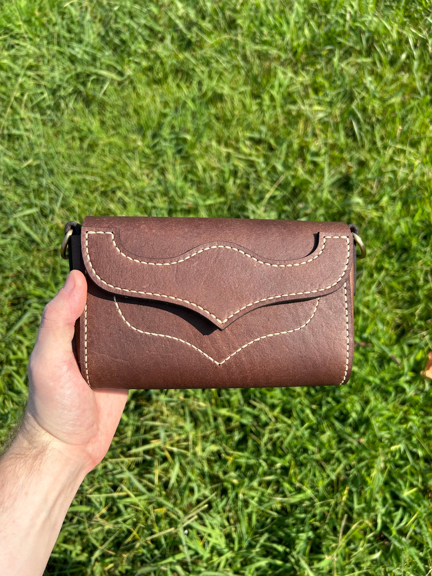 A brown leather crossbody bag with tan stitching, held in a hand against a green grass background.