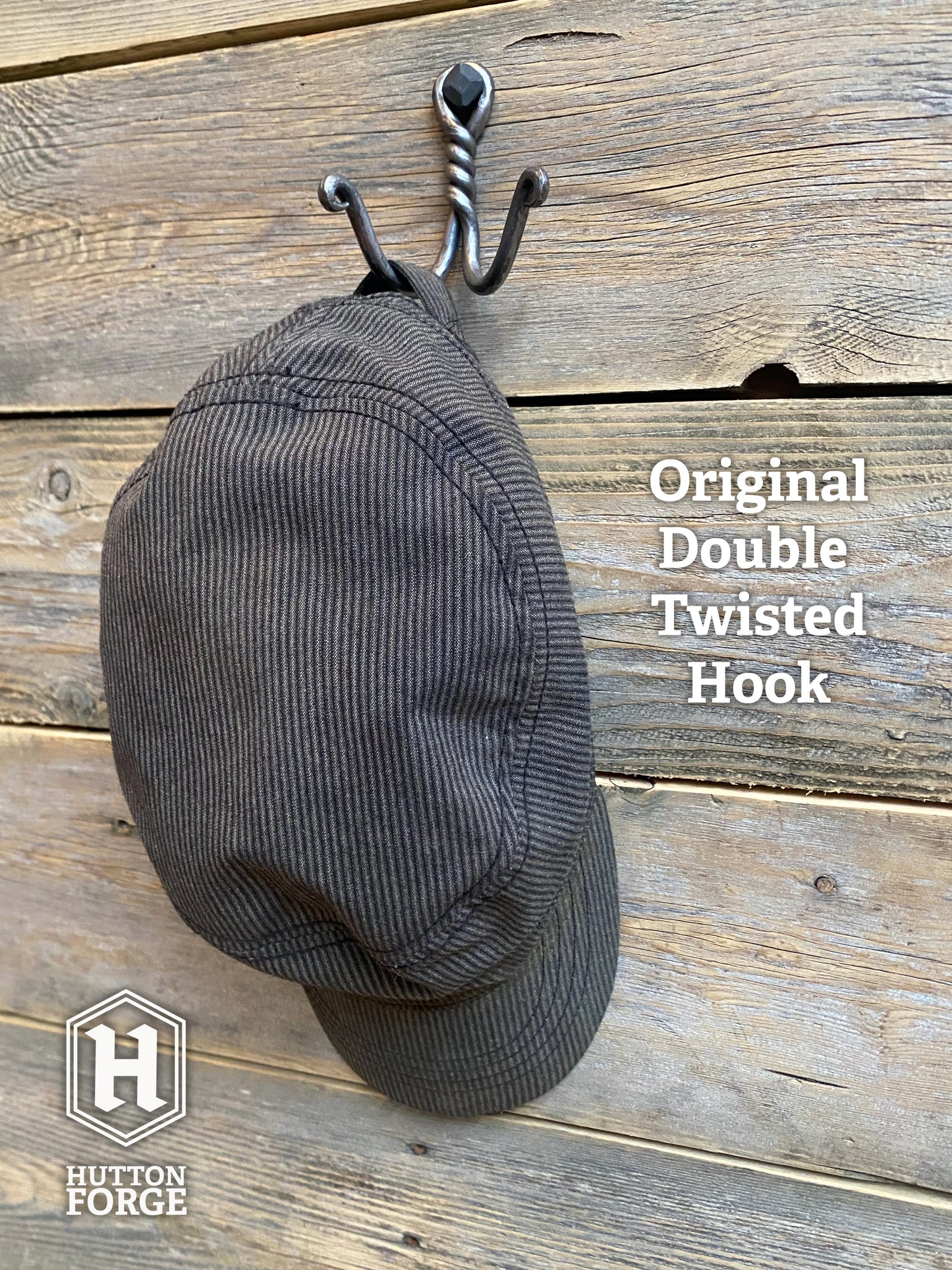 A hat hanging on a hand-forged twisted double wall hook in blackened steel, mounted with a hexagonal bolt on rustic wooden boards. Hutton Forge.
