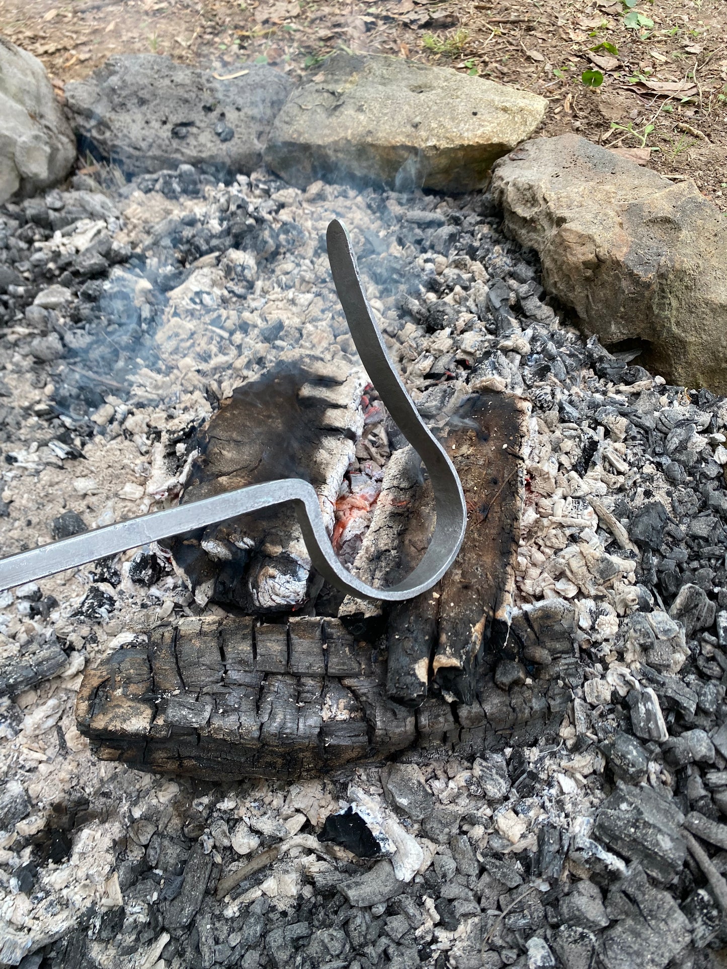 The working end of a fire poker resting over embers and burnt wood in an outdoor campfire pit with ash and rocks around it.