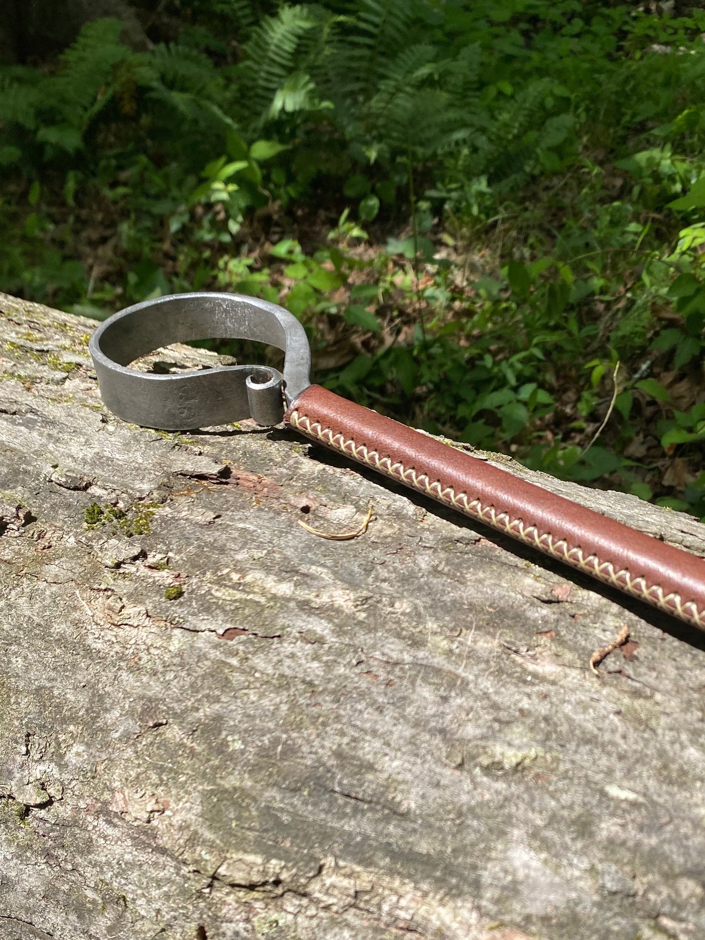 Close-up of a leather-wrapped fire poker with a hand-forged steel ring end and brown leather handle with visible stitching, lying on a weathered log surrounded by green ferns. Hutton Forge.