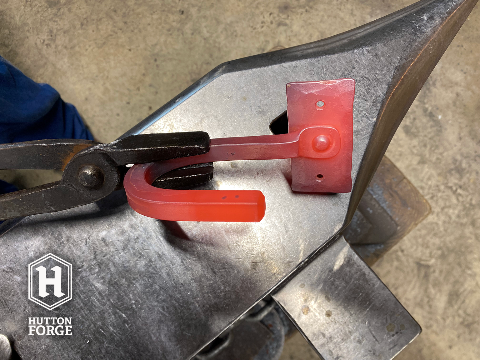 A blacksmith's forge with a glowing red metal gun hook being shaped on an anvil. The hook is held by tongs, and the Hutton Forge logo is visible in the lower left corner.
