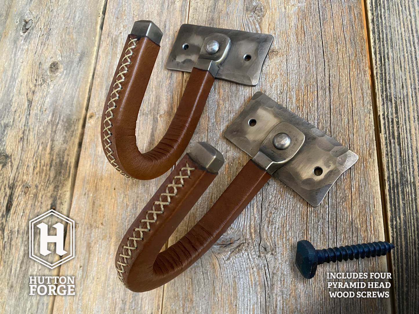 Hand-forged brushed steel J-hook wall mount wrapped in brown leather with cross-stitching, riveted to a hammered steel plate and mounted on a rustic wooden wall. Hutton Forge Brushed Steel & Brown Leather gun display hook.
