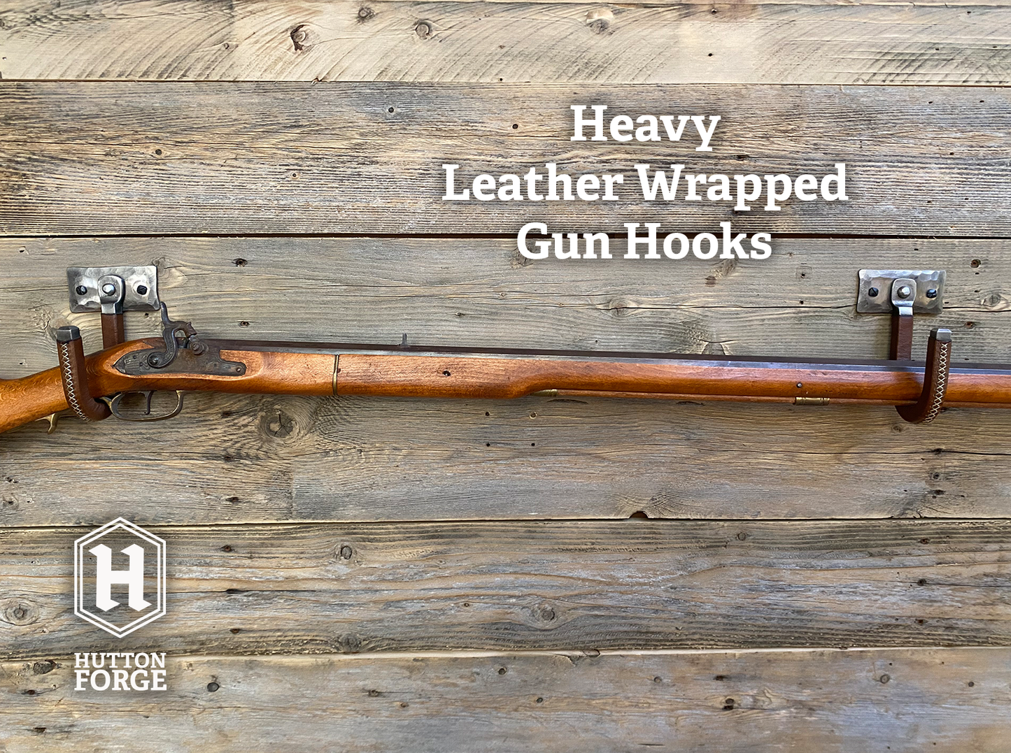 A leather-wrapped gun hook mounted on a rustic wooden plank wall, with a rifle resting horizontally across it for demonstration. Brushed Steel & Brown Leather gun display hook made by Hutton Forge.
