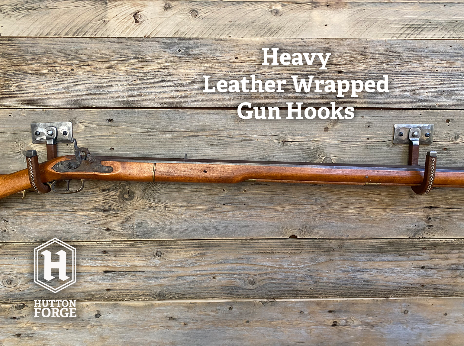 A leather-wrapped gun hook mounted on a rustic wooden plank wall, with a rifle resting horizontally across it for demonstration. Brushed Steel & Brown Leather gun display hook made by Hutton Forge.
