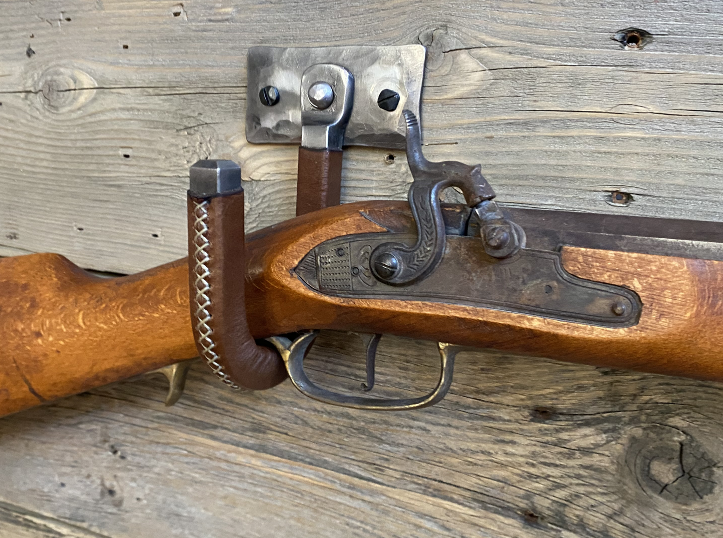 Hand-forged brushed steel J-hook wall mount wrapped in brown leather with cross-stitching, riveted to a hammered steel plate and mounted on a rustic wooden wall. Hutton Forge Brushed Steel & Brown Leather gun display hook.
