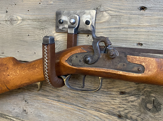 Hand-forged brushed steel J-hook wall mount wrapped in brown leather with cross-stitching, riveted to a hammered steel plate and mounted on a rustic wooden wall. Hutton Forge Brushed Steel & Brown Leather gun display hook.
