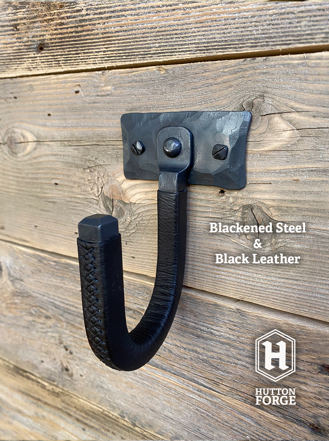 Hand-forged blackened steel gun hook wall mount wrapped in black leather, riveted to a hammered steel plate and mounted on a rustic wooden wall. Hutton Forge Blackened Steel & Black Leather gun display hook.
