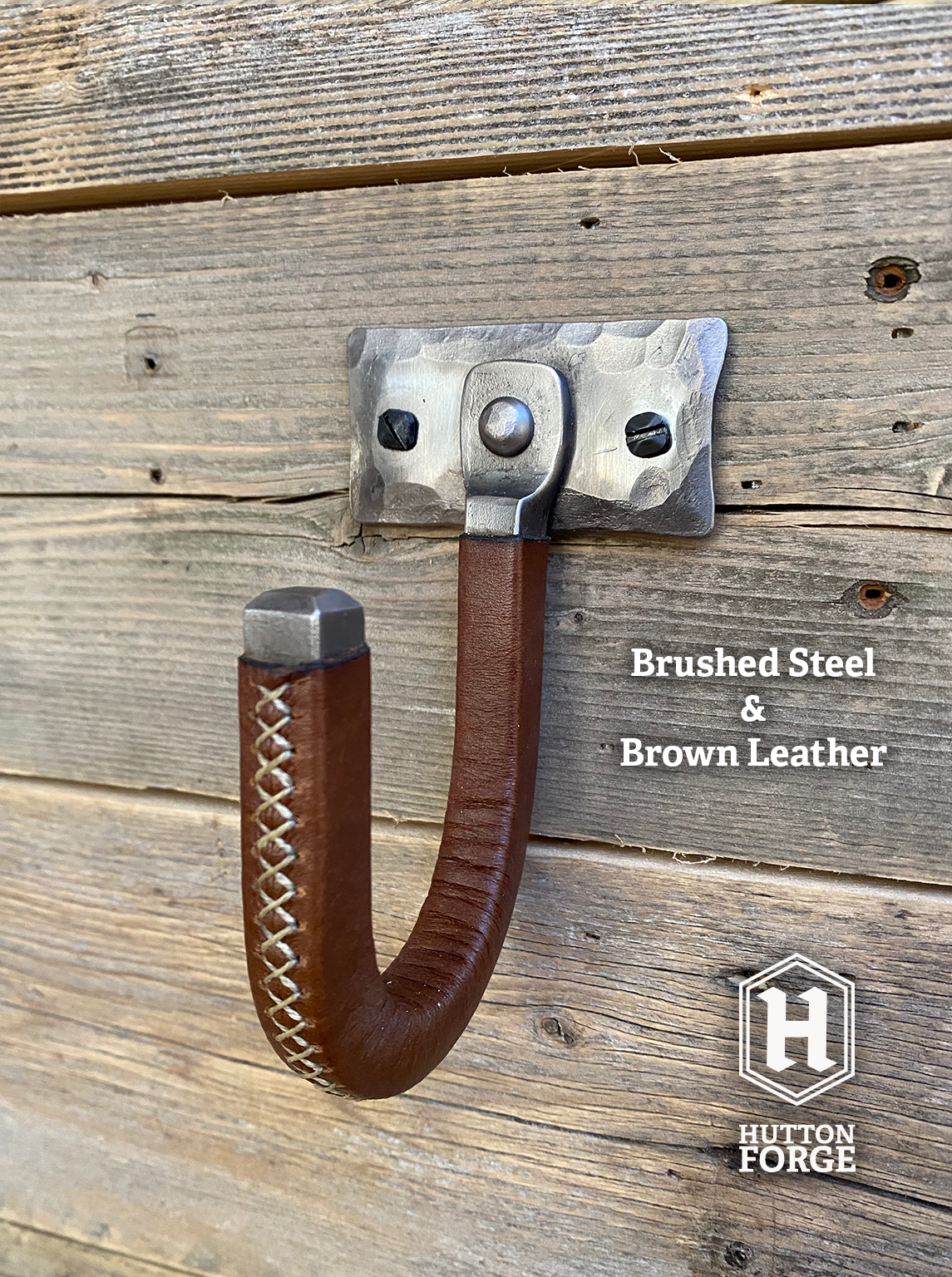 Hand-forged brushed steel gun hook wall mount wrapped in brown leather, riveted to a hammered steel plate and mounted on a rustic wooden wall. Hutton Forge Brushed Steel & Brown Leather gun display hook.
