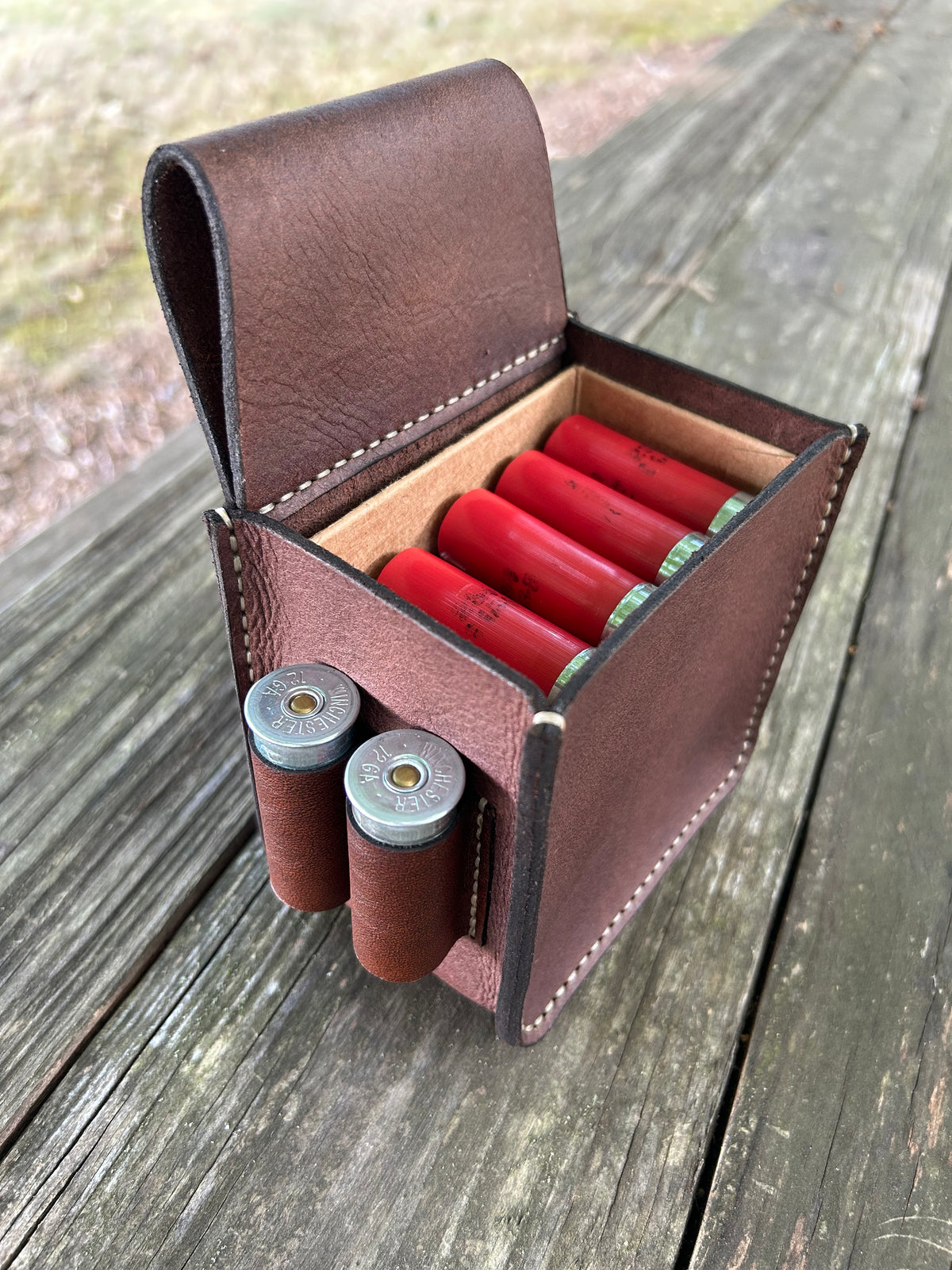 A brown leather pouch holding a shotgun ammo box, shown open on a weathered wooden table. Two shotgun shells are tucked into the front loops, and four more red shotgun shells are stored inside the padded compartment.