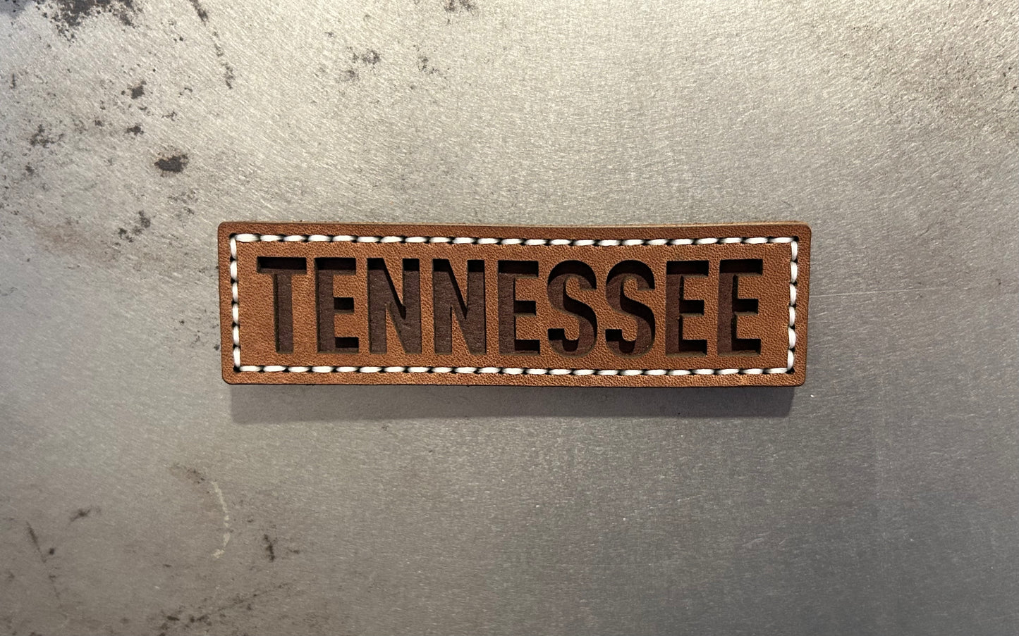 A rectangular brown leather magnet with the word TENNESSEE in dark brown letters, bordered by white stitching. Hutton Forge. 