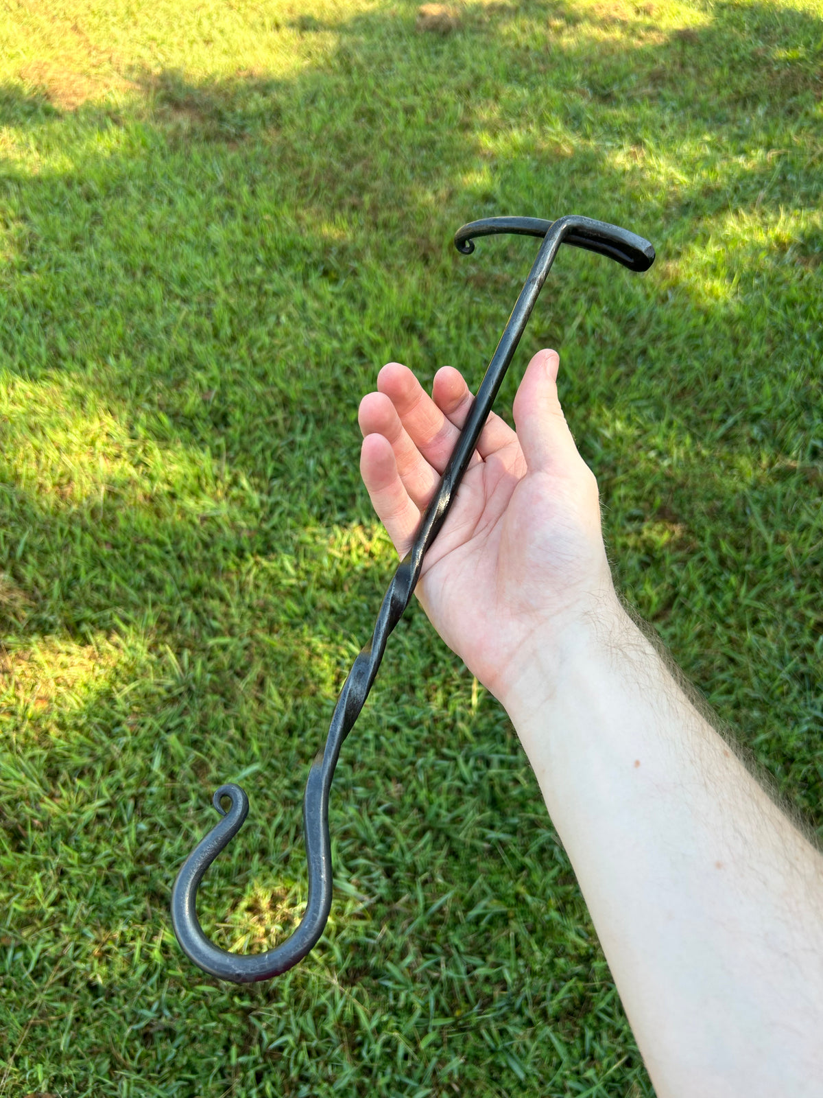 Blacksmith-made Dutch oven lid lifter with a twisted steel shaft, hooked end, and curved handle, held in a hand for scale. Hutton Forge.