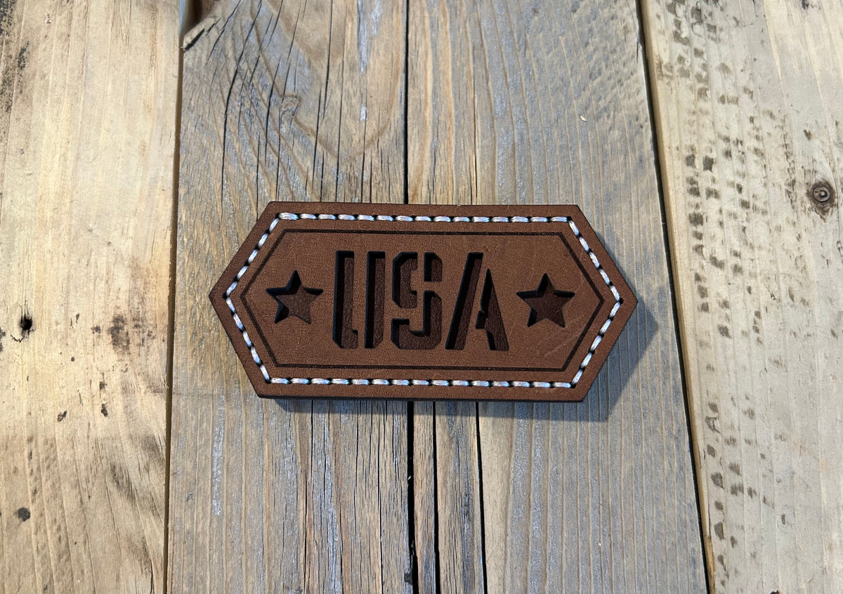 Hand-stitched brown leather magnet with cut-out “USA” lettering and stars, featuring white stitching on a hexagonal design. Hutton Forge.
