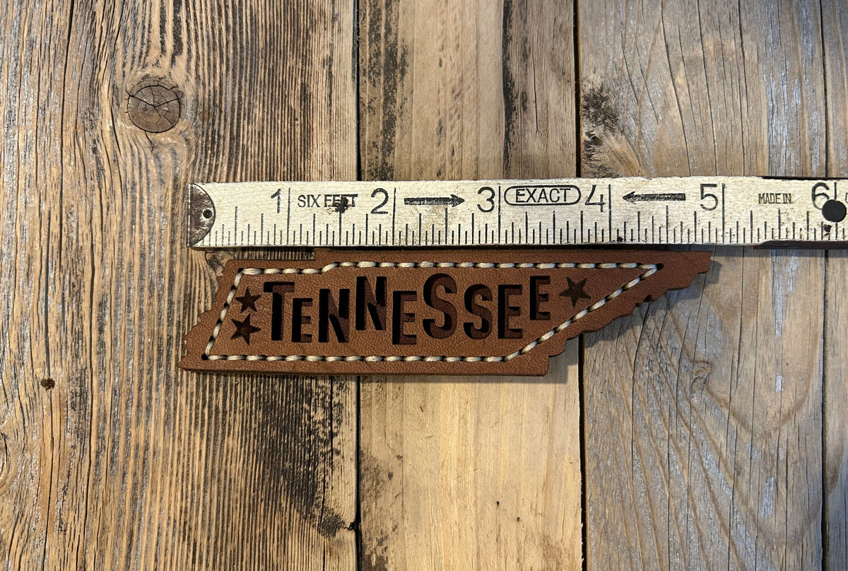 A brown leather cutout of the state of Tennessee, with the state's name cut out in the center. The word "Tennessee" is visible, along with three star cutouts. The edges are stitched with tan thread. Hutton Forge.
