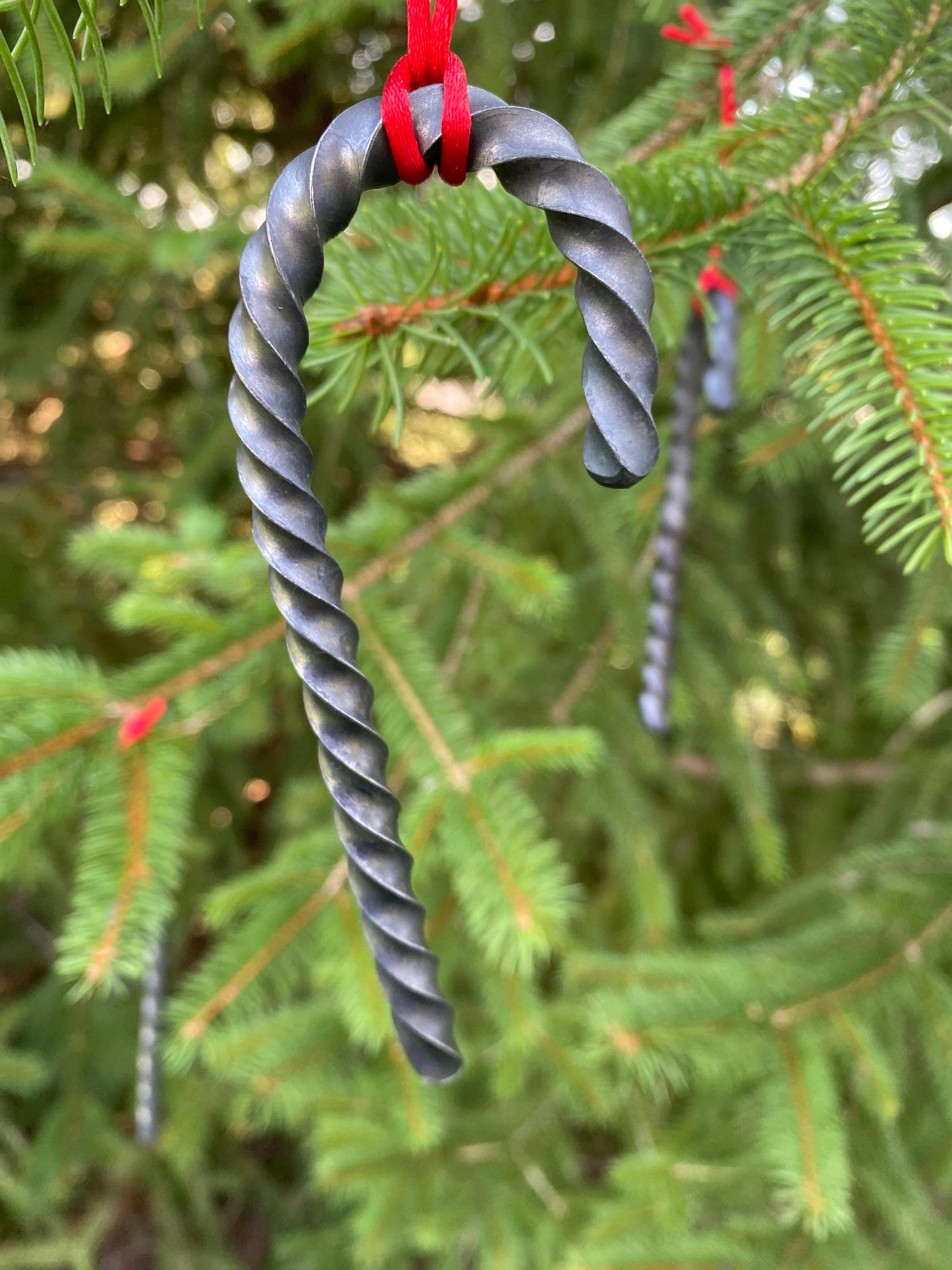  A Hand Forged Twisted Steel Candy Cane Ornament hanging from a red cord on a tree. Hutton Forge.