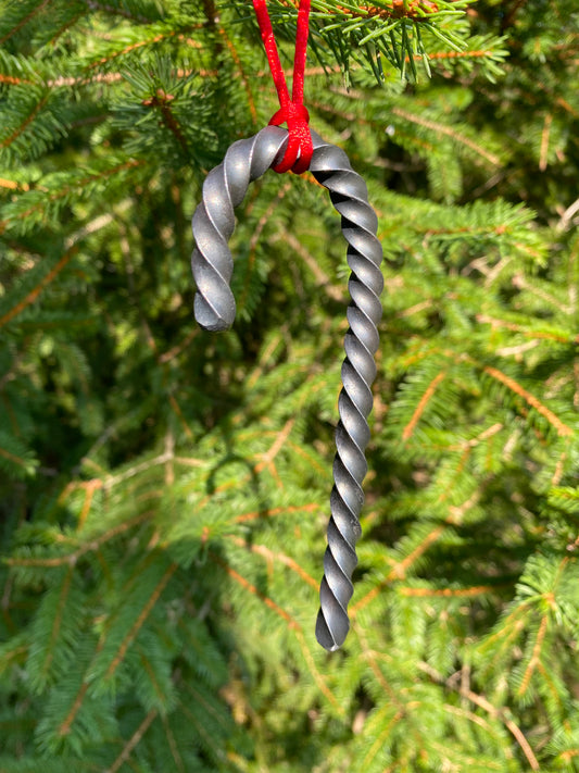 A twisted metal candy cane ornament hanging from a red string against a blurred background of green evergreen branches. Hutton Forge