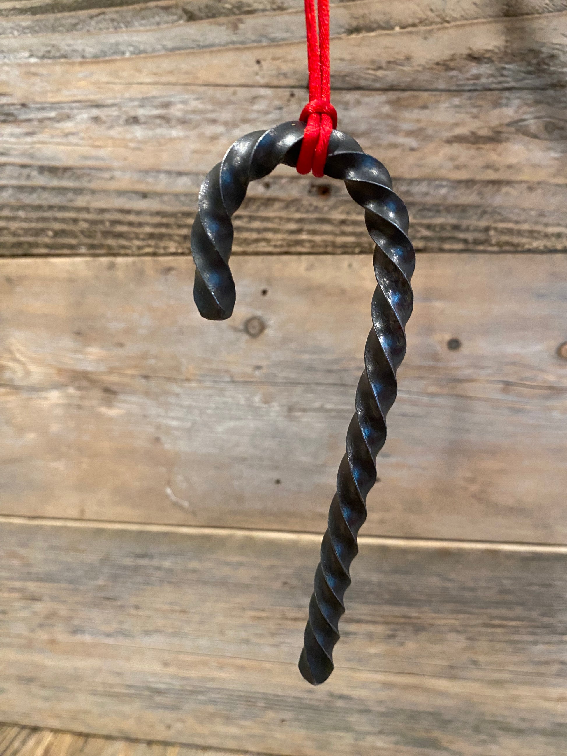  A Hand Forged Twisted Steel Candy Cane Ornament hanging from a red cord against a rustic wooden plank background. Hutton Forge.
