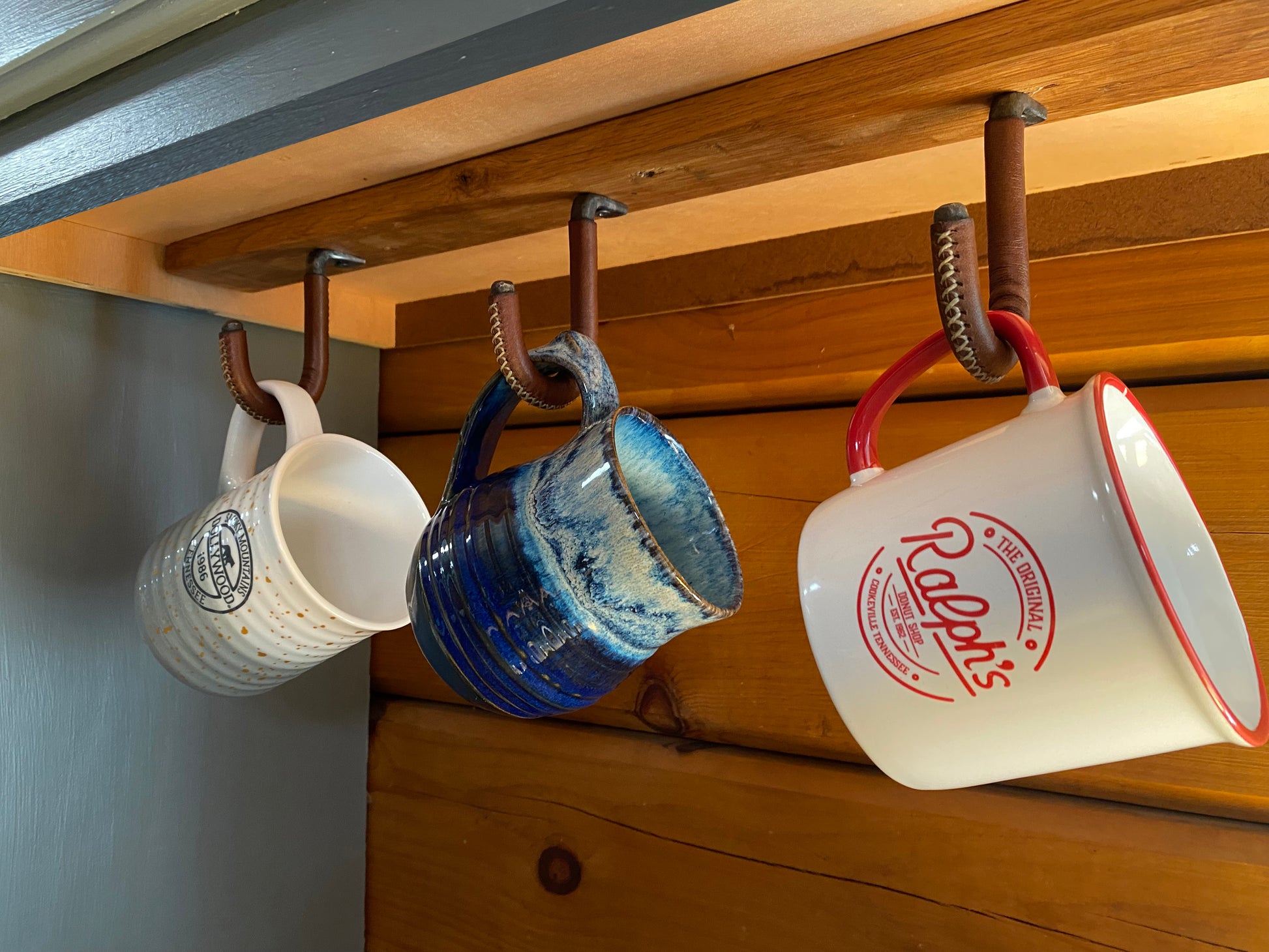 Use multiple hooks to create a unique under cabinet display of your favorite coffee mugs. Three ceramic mugs hanging from rustic leather wrapped metal hooks under kitchen cabinets.  