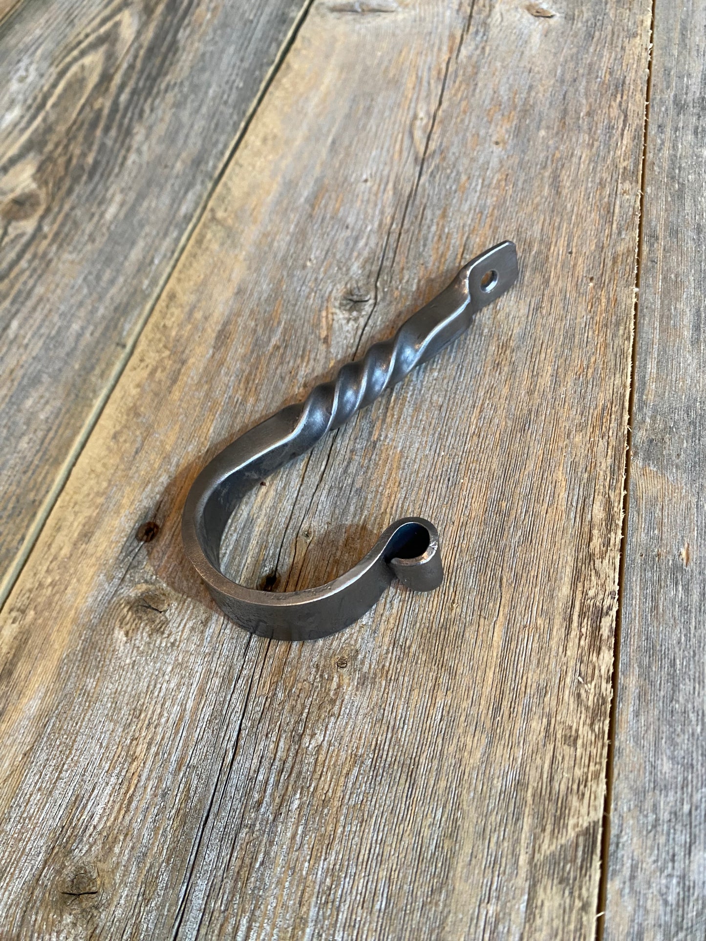 Hand-forged twisted steel J-hook in blackened finish, blacksmith-made with a decorative twist, shown on a rustic wooden surface. Hutton Forge.

