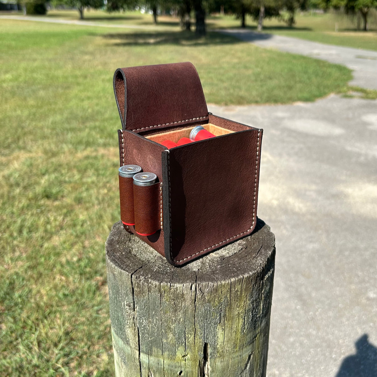 A brown leather shotgun case, shown on a wooden post outdoors. Two shotgun shells are tucked into the side pockets, and a red shotgun shells are inside.