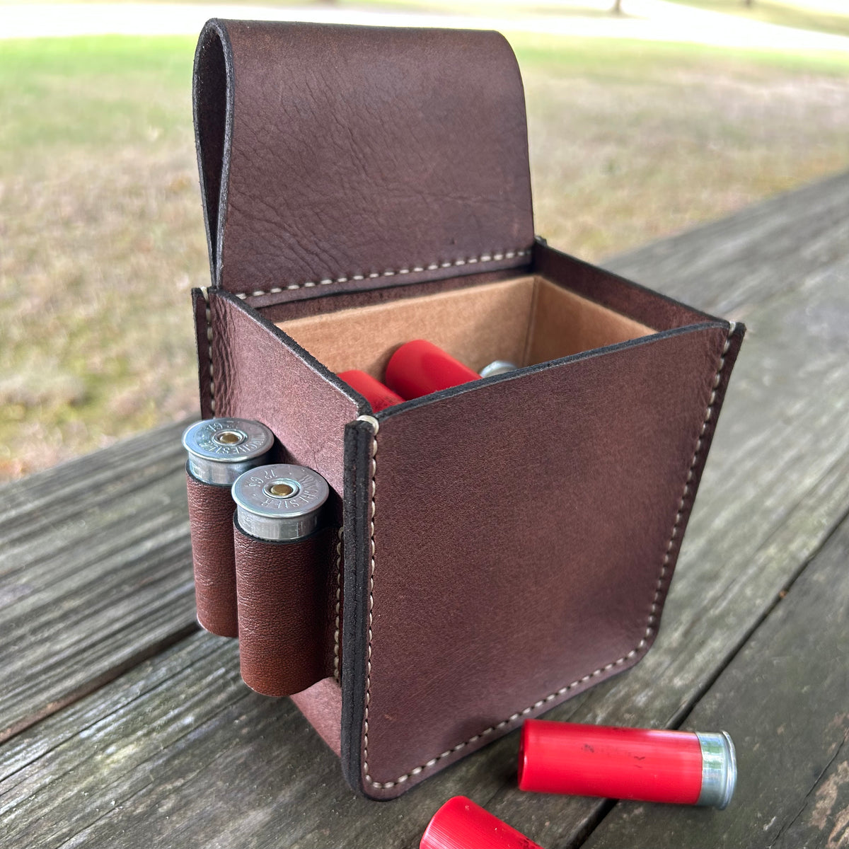 A brown leather shotgun shell holder pouch, shown open on a weathered wooden table with shotgun shells visible inside and one lying outside.