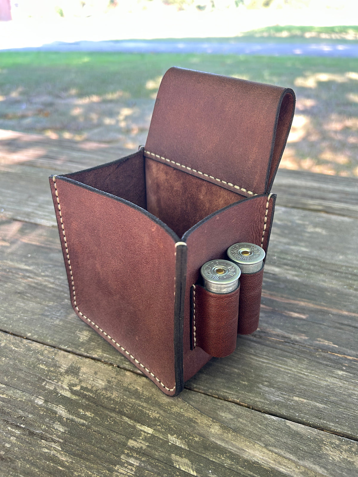 A empty brown leather shotgun shell holder pouch with white contrast stitching, shown open on a weathered wooden table with two shotgun shells in the side pockets, set against an outdoor blurred background.