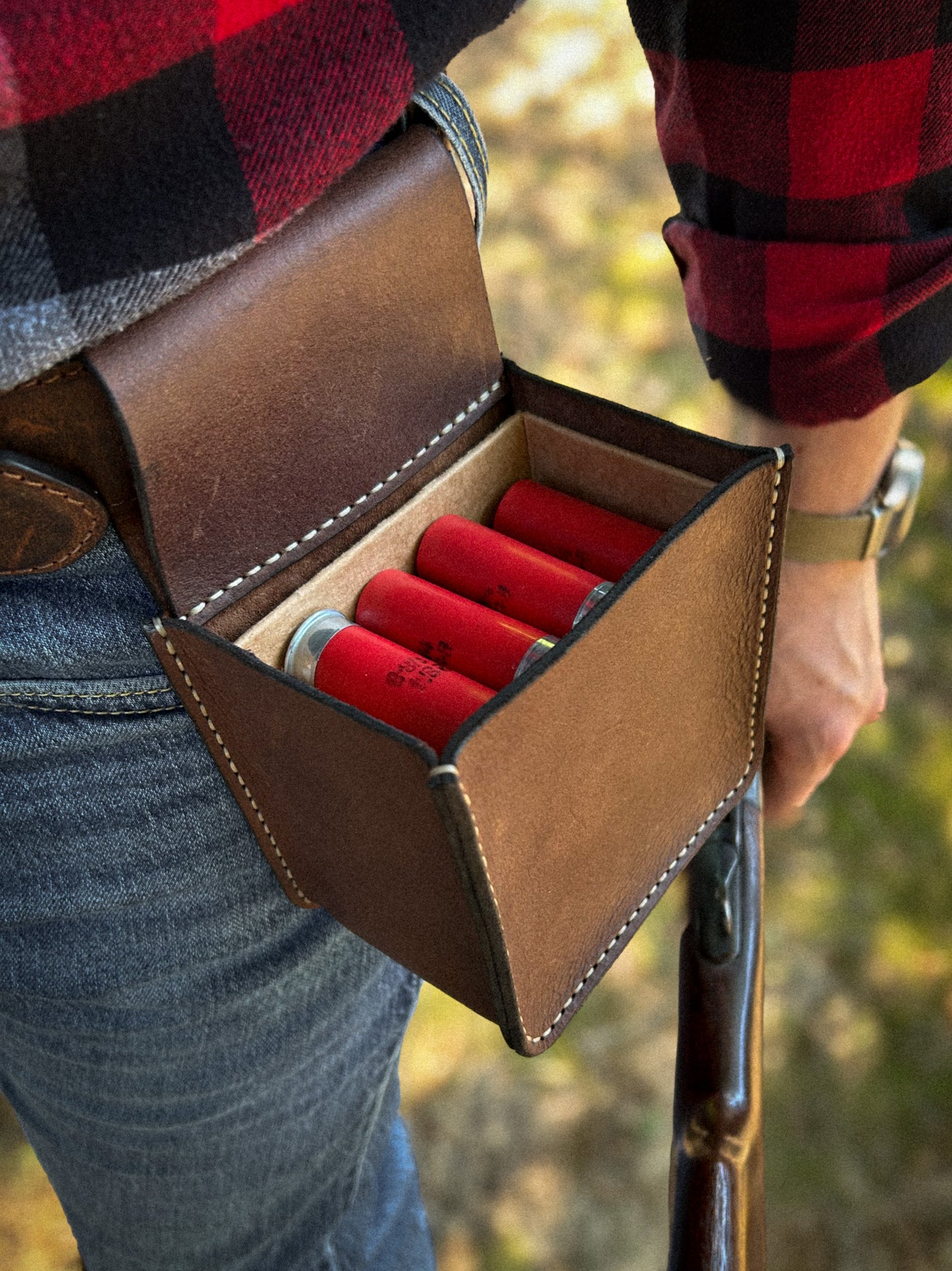 A brown leather shotgun shell pouch worn on a person's hip, holding red shotgun shells, with a red and black plaid shirt and blue jeans visible against an outdoor blurred background.