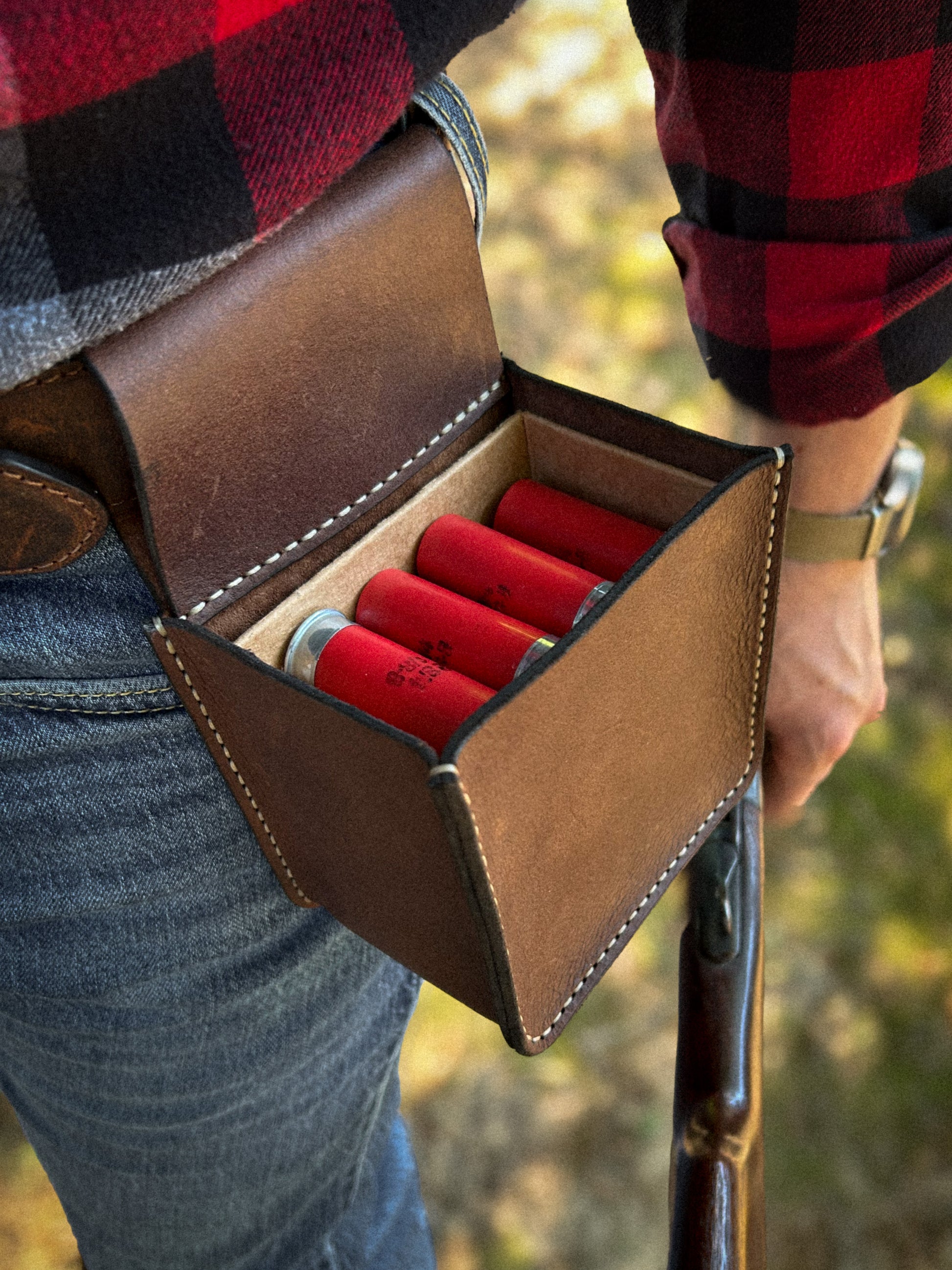 A brown leather shotgun shell pouch worn on a person's hip, holding red shotgun shells, with a red and black plaid shirt and blue jeans visible against an outdoor blurred background.
