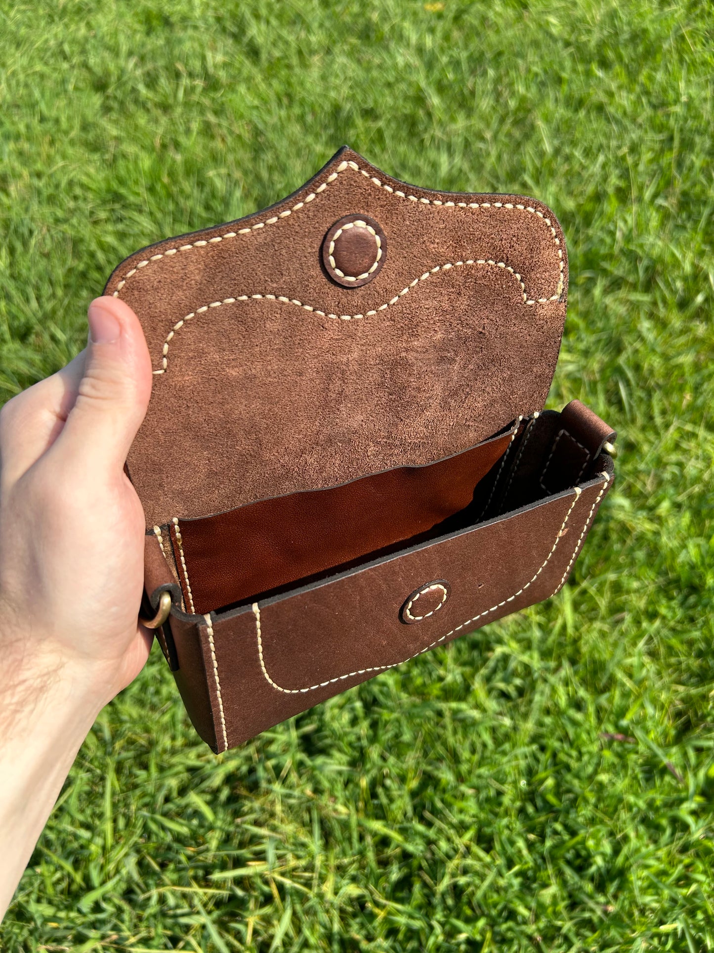A brown leather purse held open by a hand against a green grass background. The pouch features tan stitching, a magnetic button closure, and a visible brown interior.