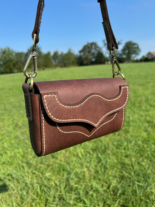 A brown leather crossbody bag with a flap closure, decorative stitching, and a western flap detail, hanging outdoors against a blurred green field and blue sky background.