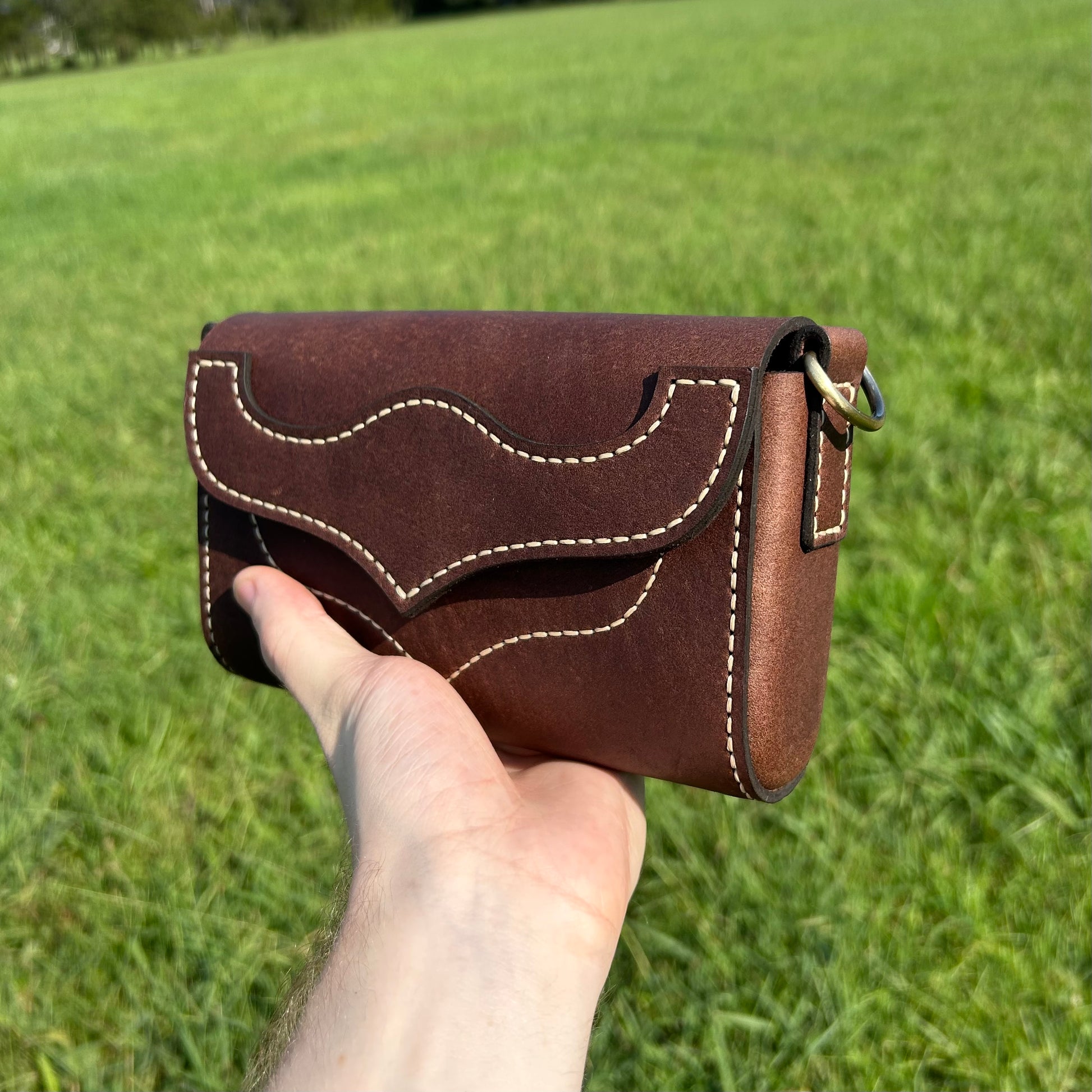 A hand holding a small brown leather purse with tan stitching, set against a blurred green grass background.
