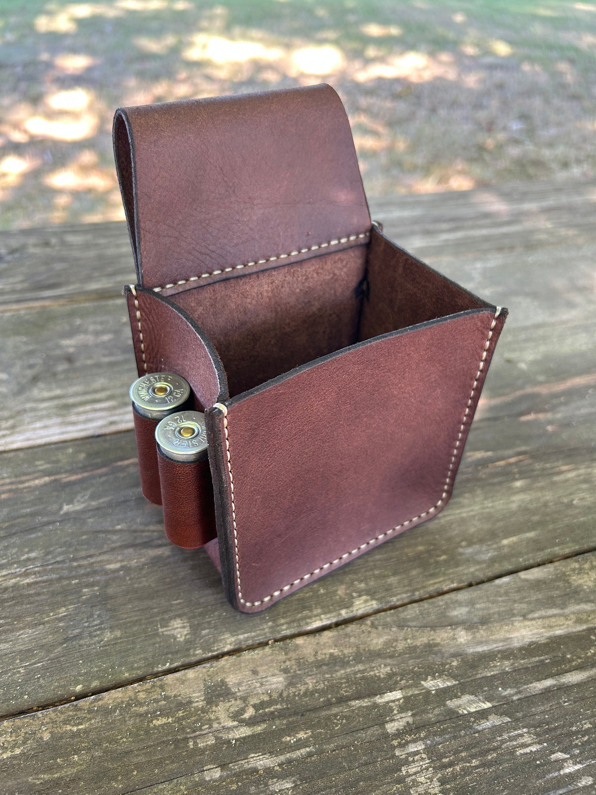 A brown leather shotgun shell holder pouch with white stitching, shown open on a rustic wooden deck with two shotgun shells inserted.