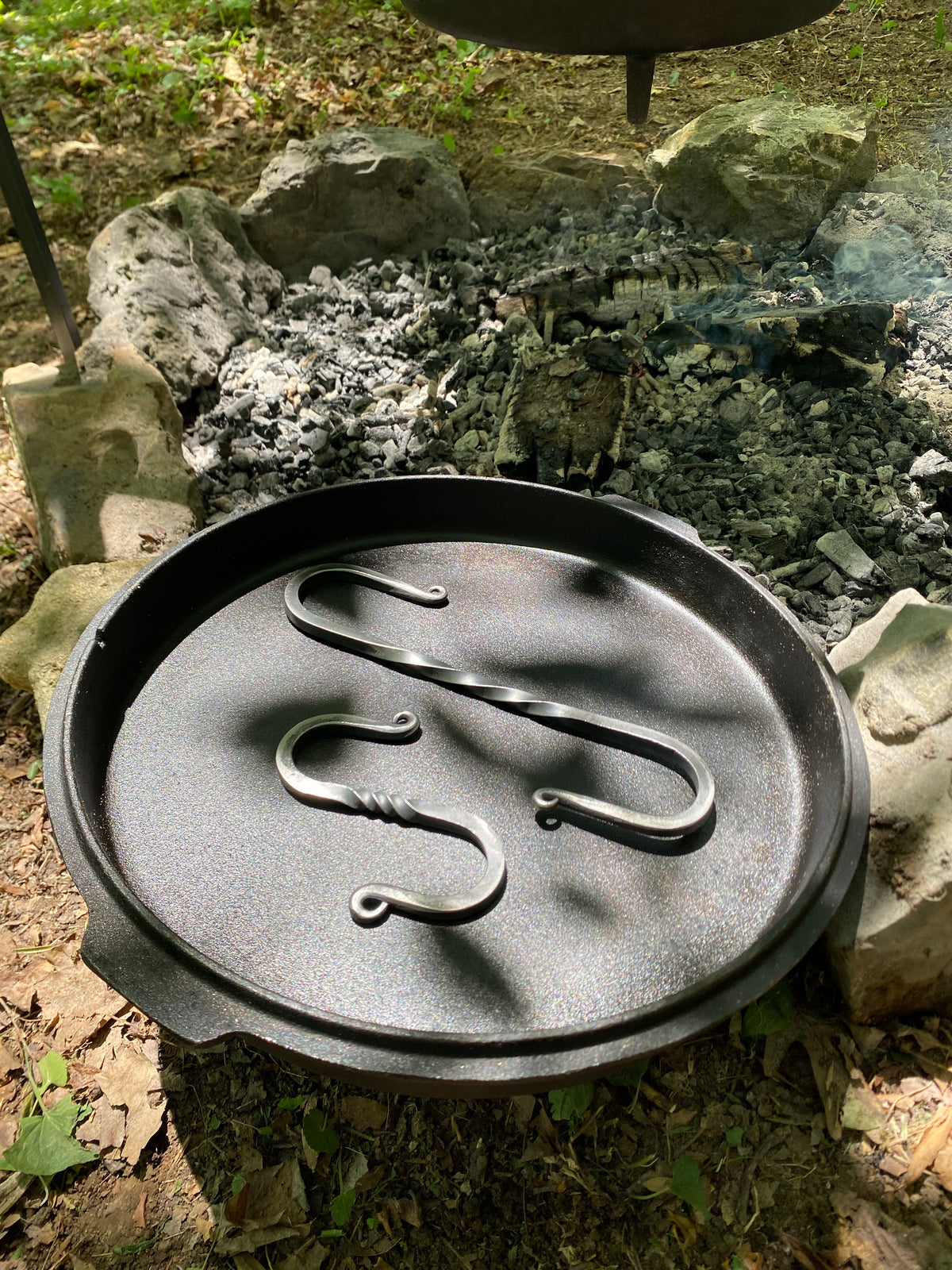 A black cast iron Dutch oven lid with two metal S-hooks, small and large, photographed outdoors on a campfire stone ring.