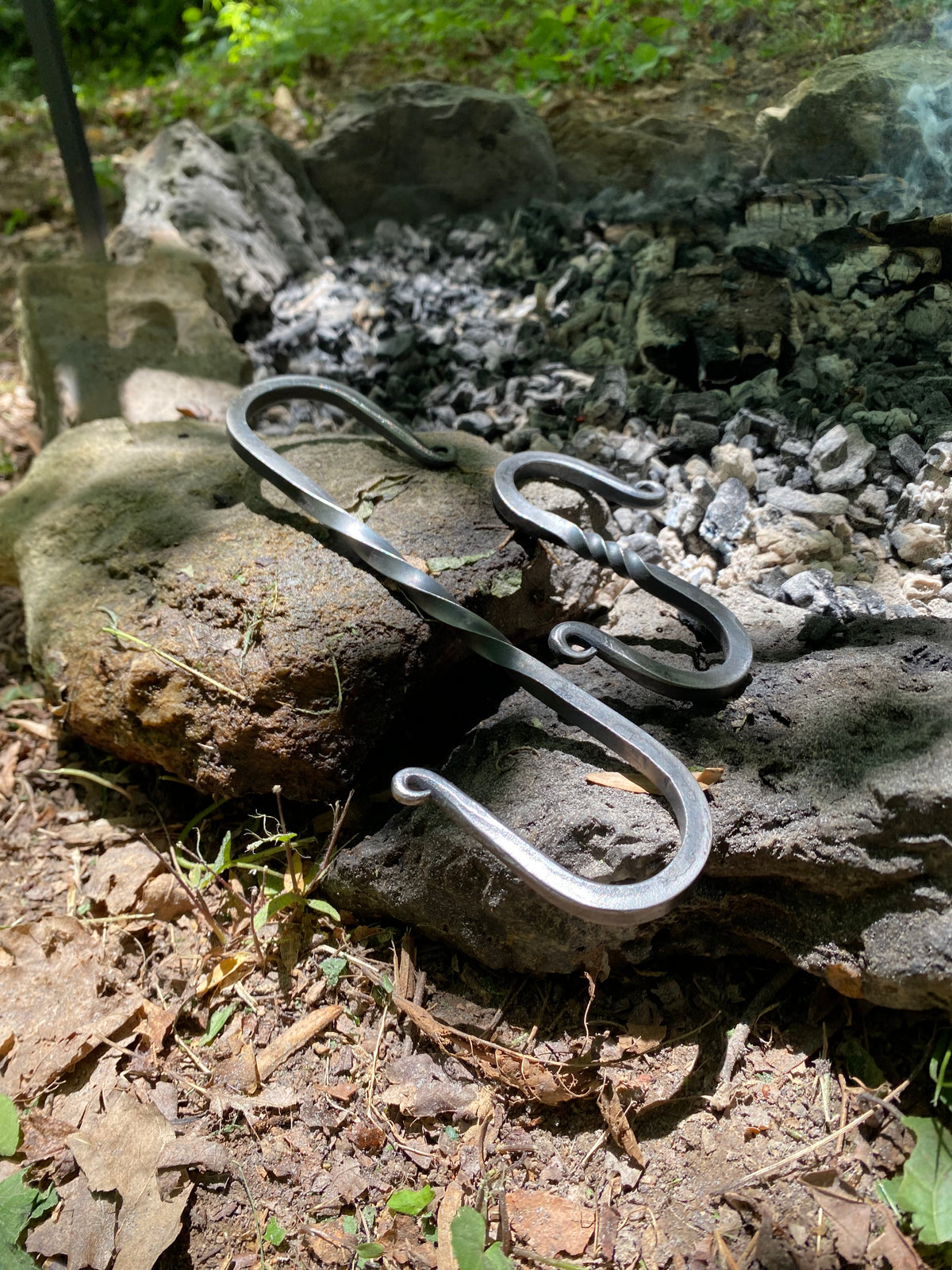 A pair of dark metal S-hooks, small and large,  placed on rough outdoor rocks. Outdoor Cooking with Hutton Forge. 