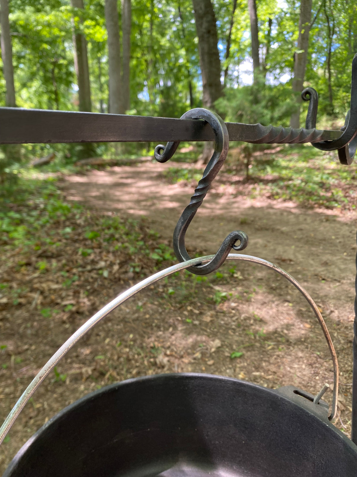  A handcrafted, dark metal campfire cooking set with an S-hook and a black cast iron pot. The hook can be used to hang the pot over a fire. Hutton Forge.