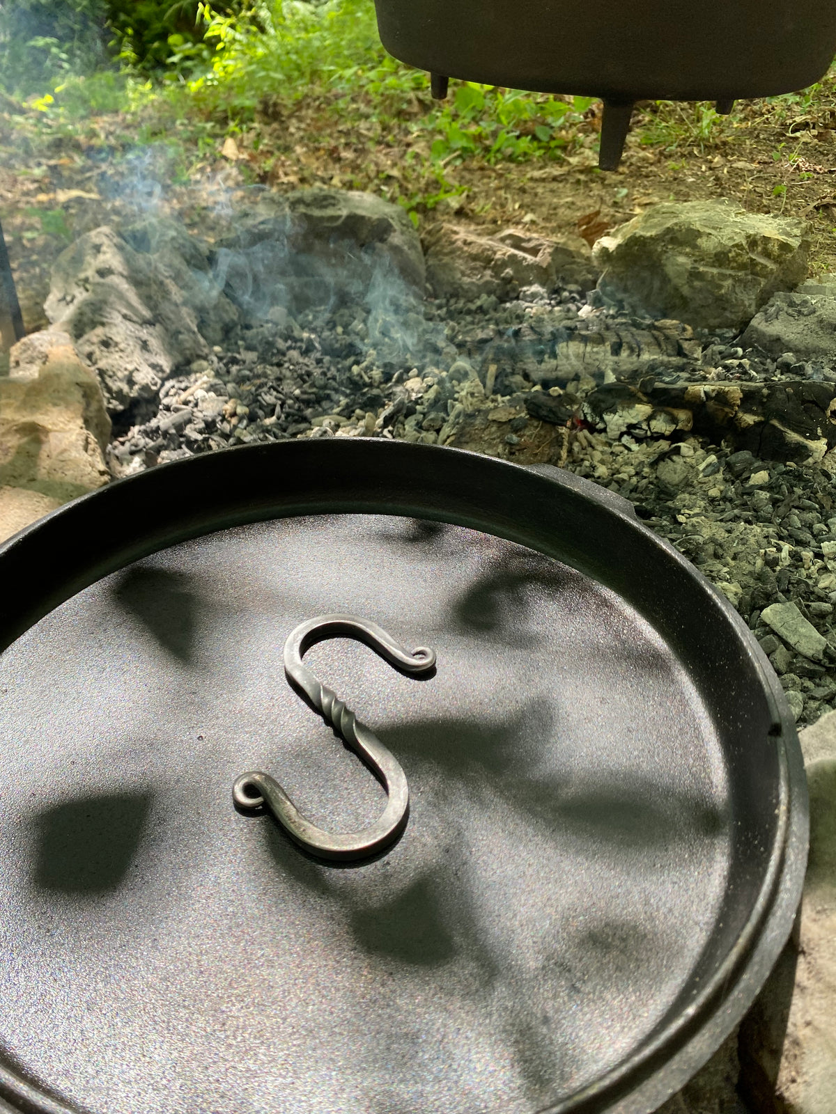 A close-up of a dark cast iron Dutch oven lid with a twisted S hook, ready to assist in cooking over the fire. 