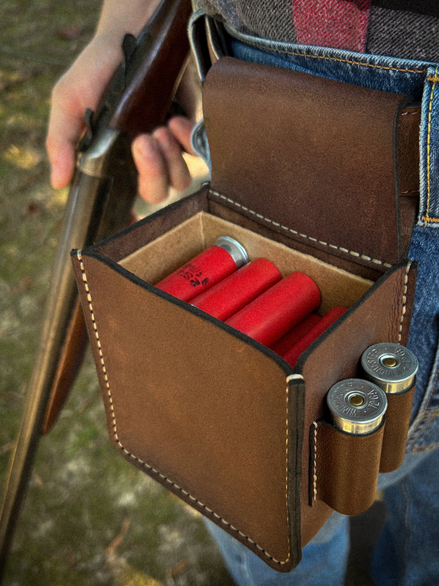 A brown leather bag that holds a box of 12 gauge shotgun shells. The person is holding a vintage firearm.