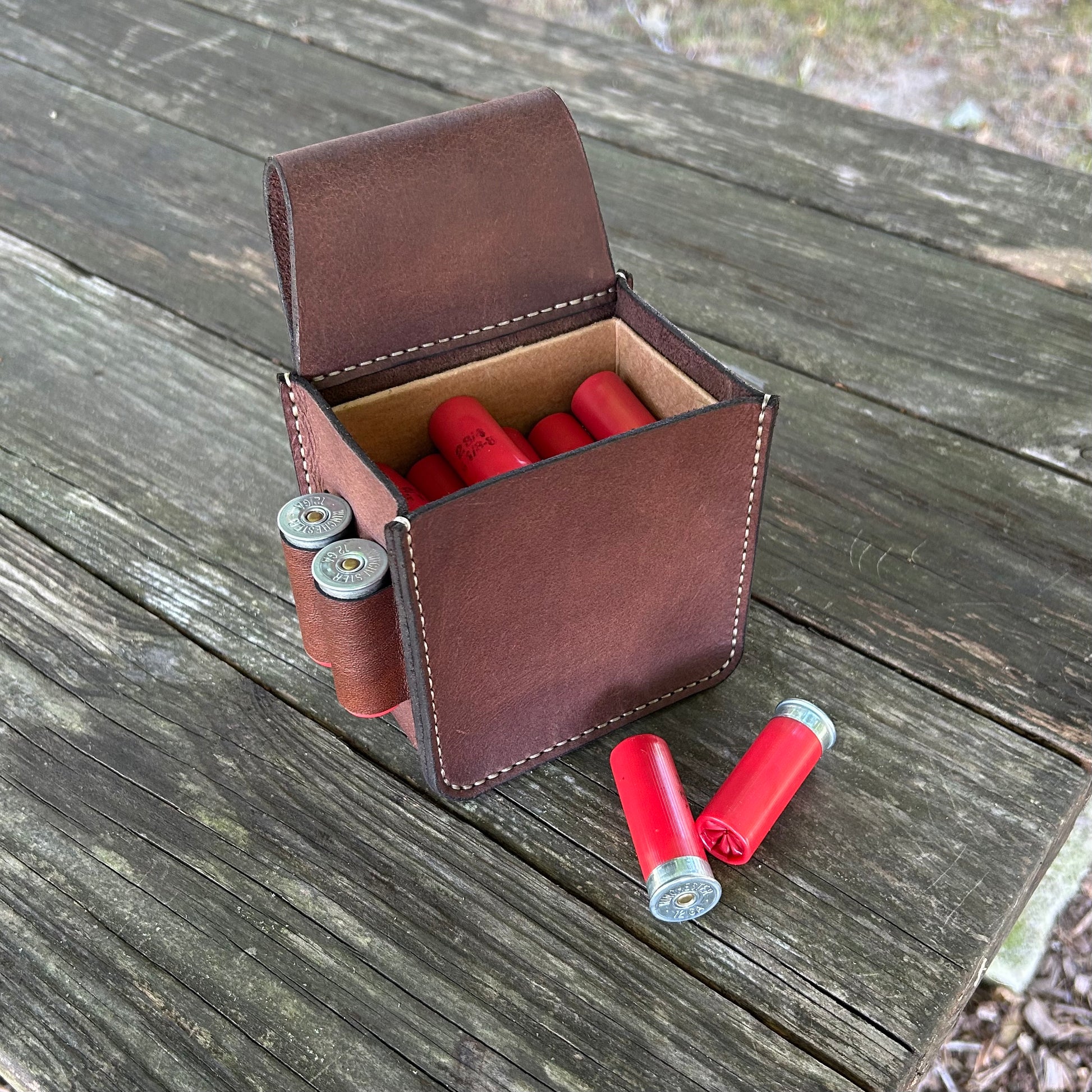 A brown leather shotgun ammo pouch, shown open on a weathered wooden table. Several red shotgun shells are visible inside the case, and two more red shells lie outside on the table.