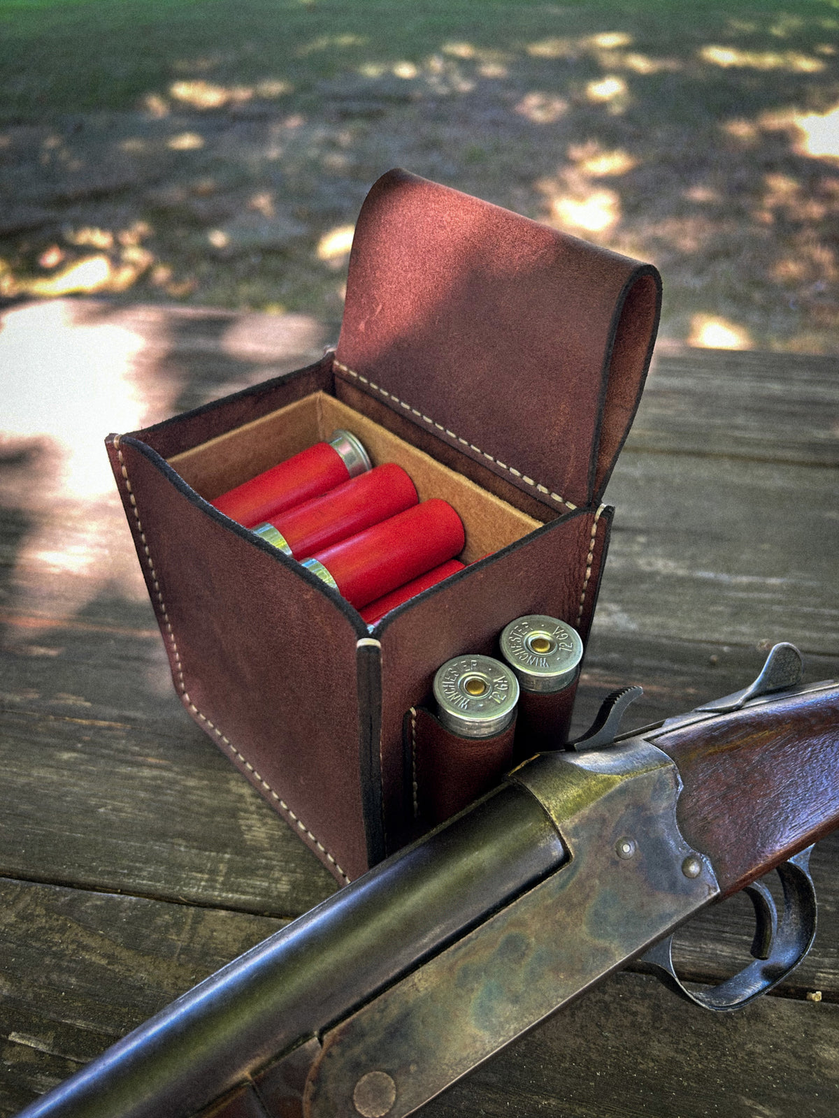 A brown leather shotgun shell case holding red shotgun shells, placed on a wooden table outdoors with part of a shotgun visible in the foreground.