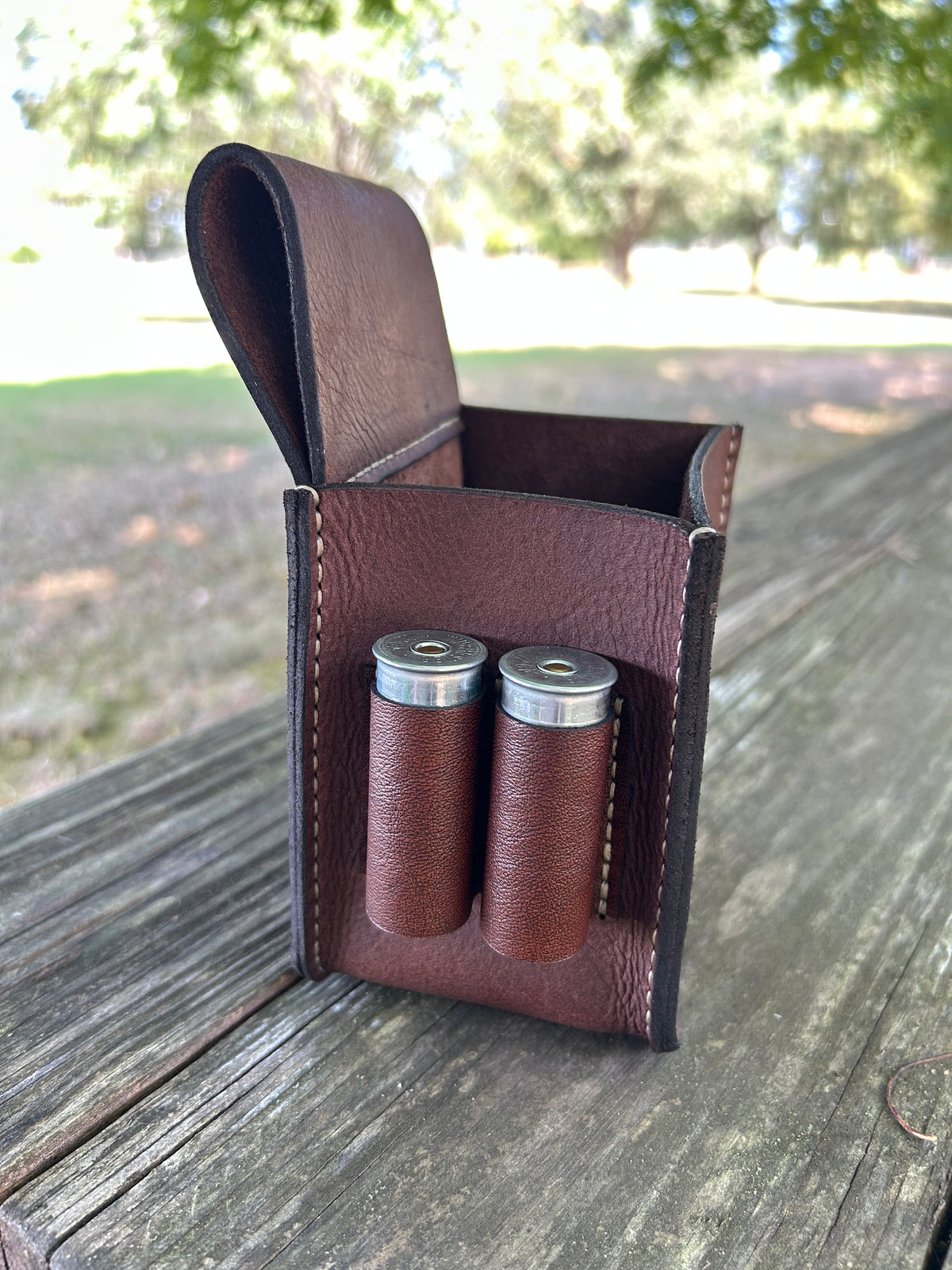 A brown leather shotgun case with two loops holding shells, shown open on a weathered wooden table with a blurred outdoor background of trees and grass.