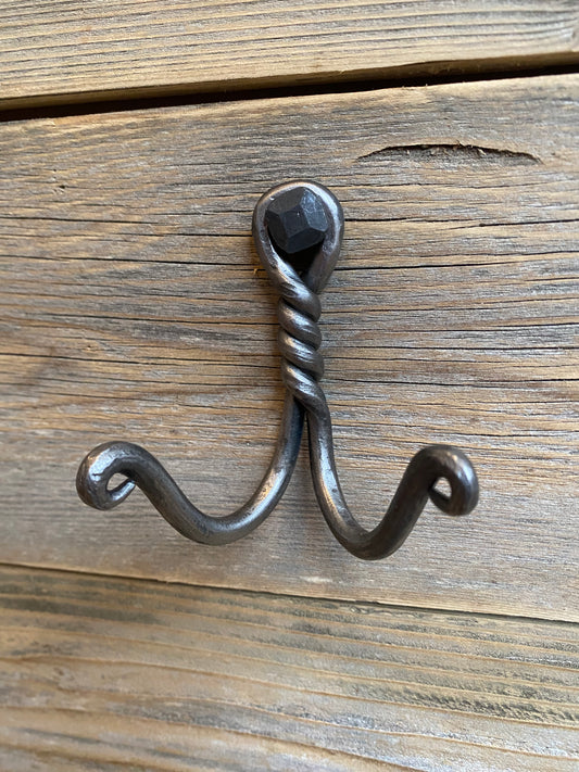 Hand-forged twisted double wall hook in blackened steel, mounted with a hexagonal bolt on rustic wooden boards. Hutton Forge.

