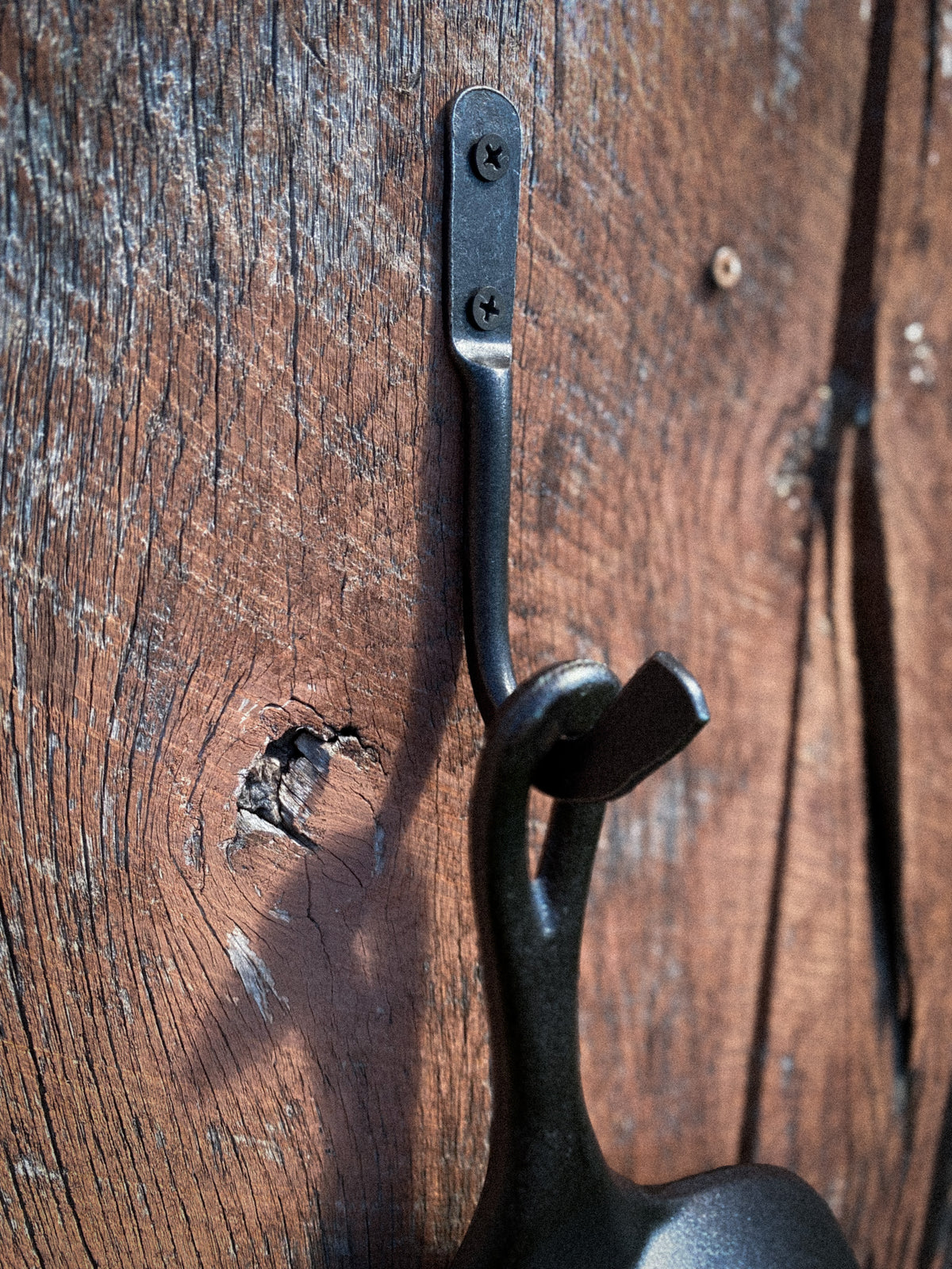 Hand-forged steel skillet hook holding a Lodge cast iron pan by the helper handle on a weathered wood wall. Hutton Forge.
