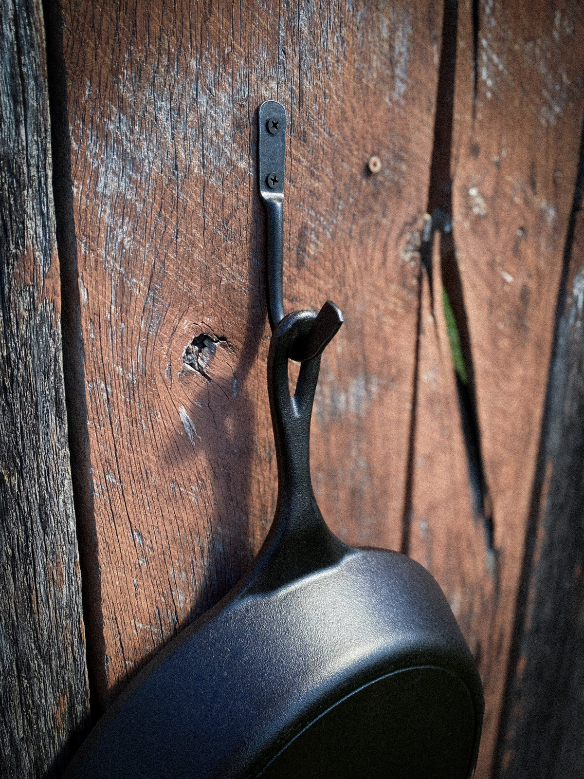 Hand-forged steel skillet hook holding a Lodge cast iron pan by the helper handle on a weathered wood wall. Hutton Forge.
