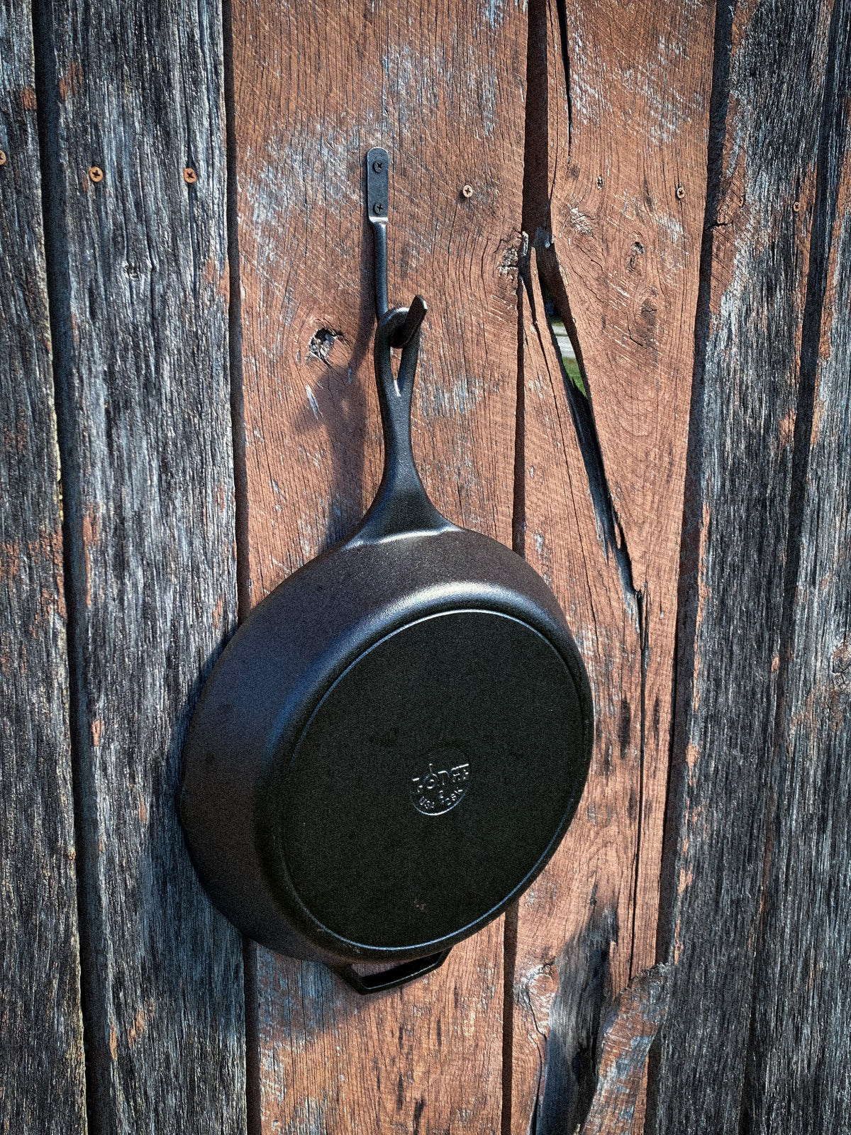 Hand-forged steel skillet hook holding a Lodge cast iron pan by the helper handle on a weathered wood wall. Hutton Forge.

